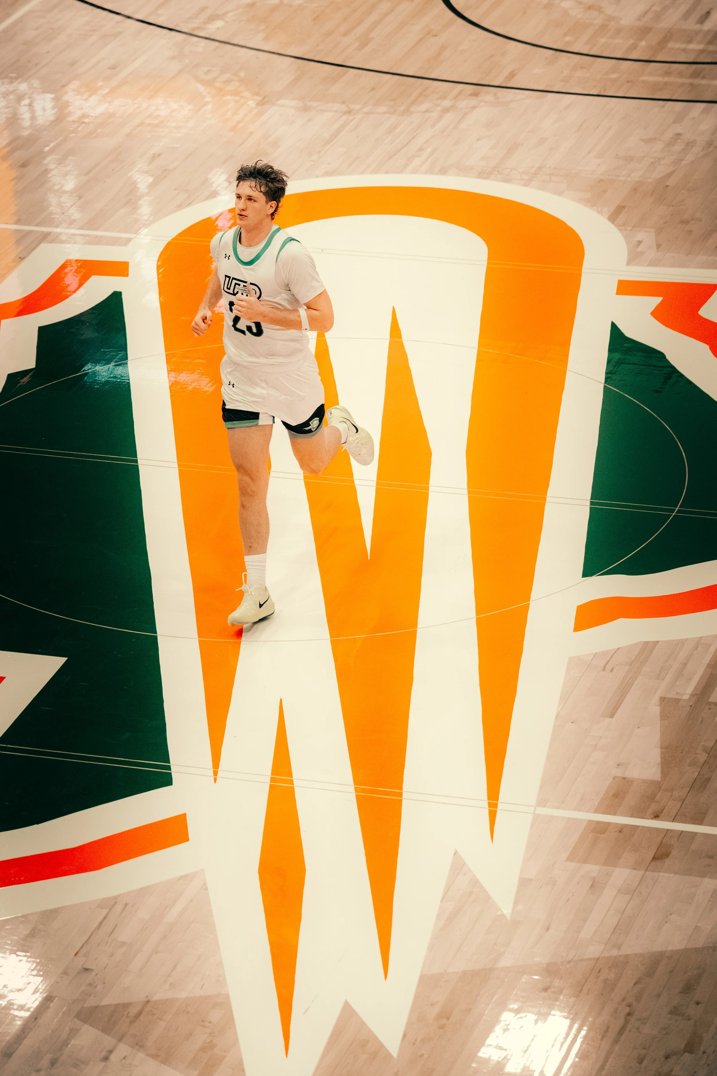 A male basketball player running on a basketball court with an orange and white logo and green accents on the floor.