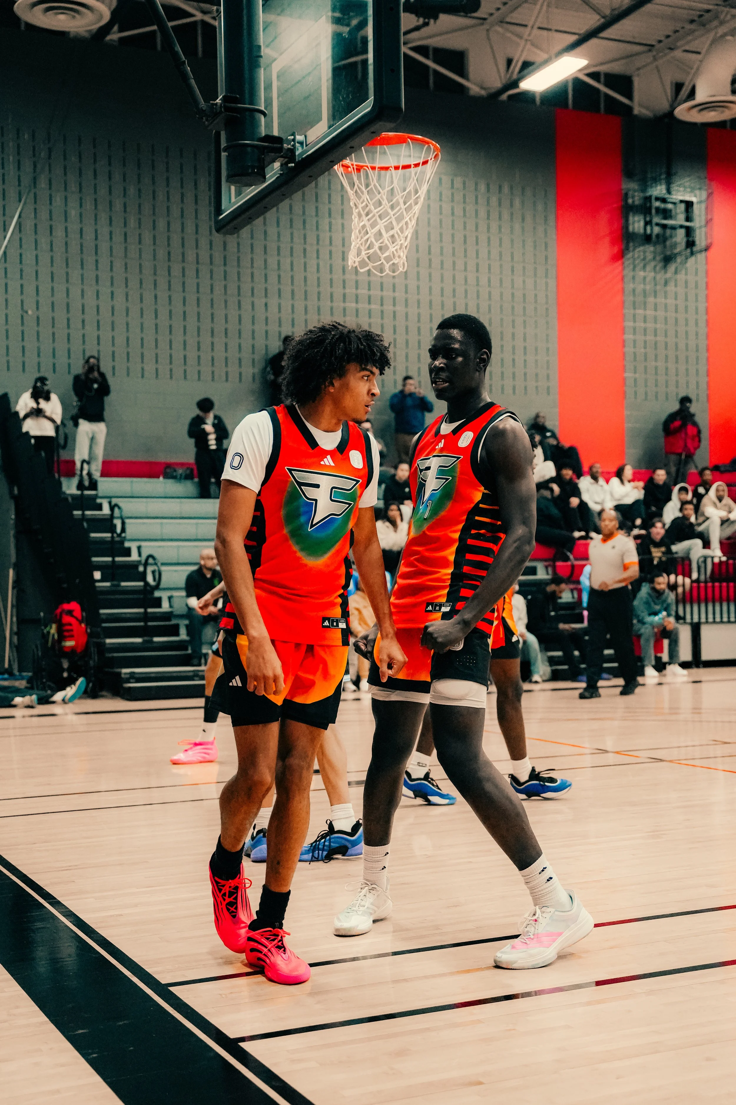 Two male basketball players in colorful jerseys standing near a basketball hoop on an indoor court, with spectators in the background.