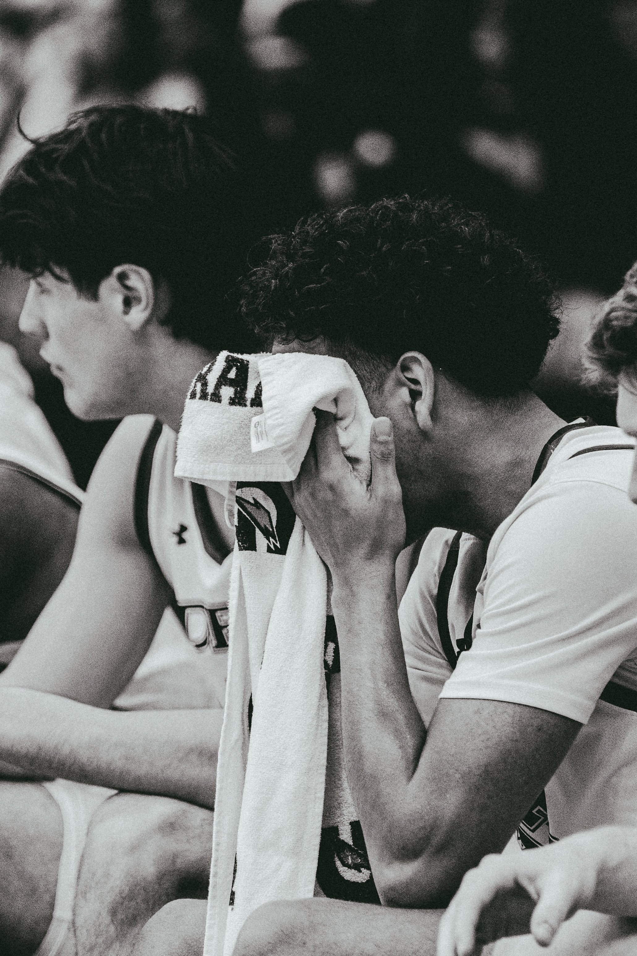 Two young men sitting side by side, one covering his face with a towel, in a black-and-white sports setting.