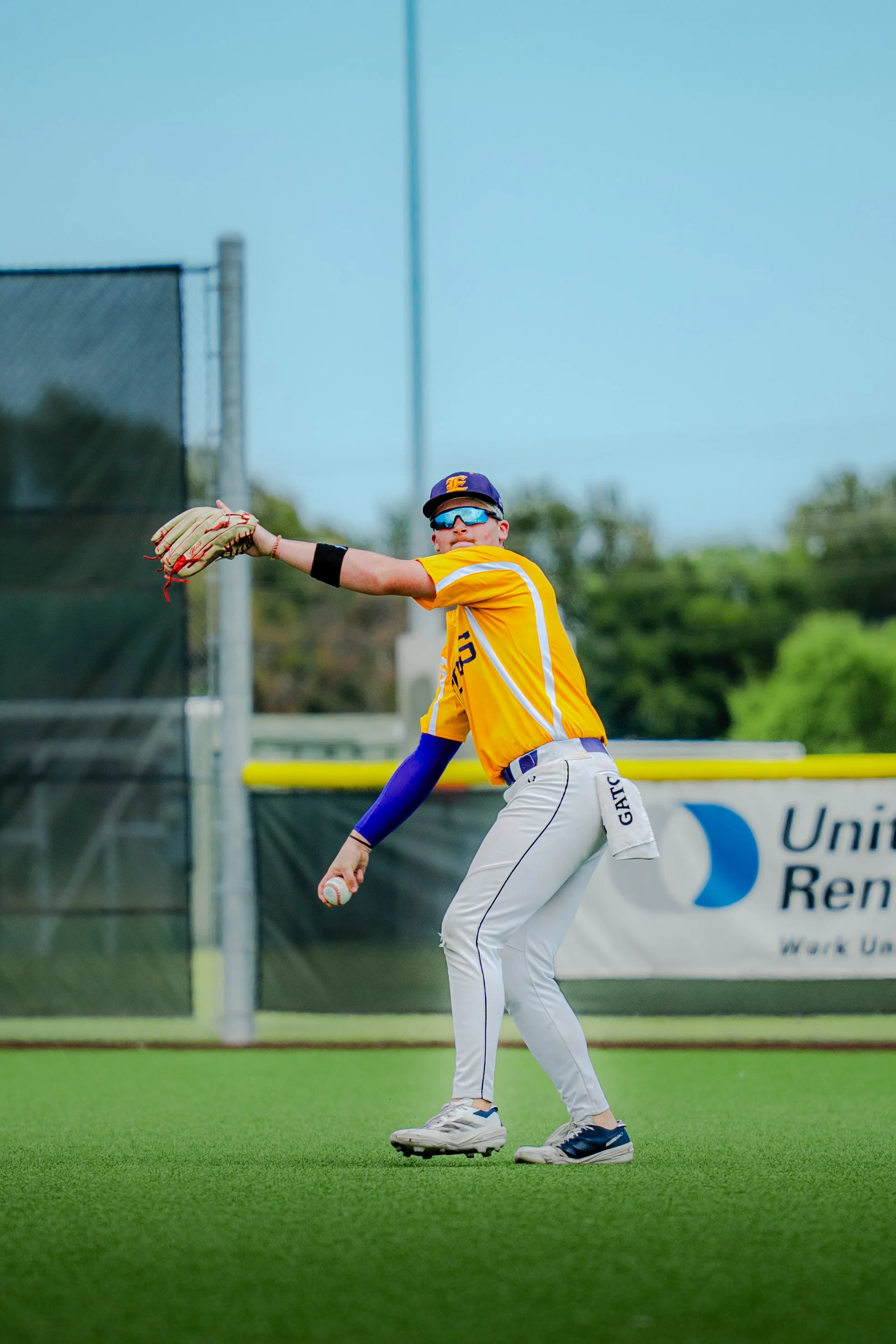A young baseball player in yellow jersey and white pants standing on the field, catching a ball with his glove, on a grassy field with a fence and trees in the background.