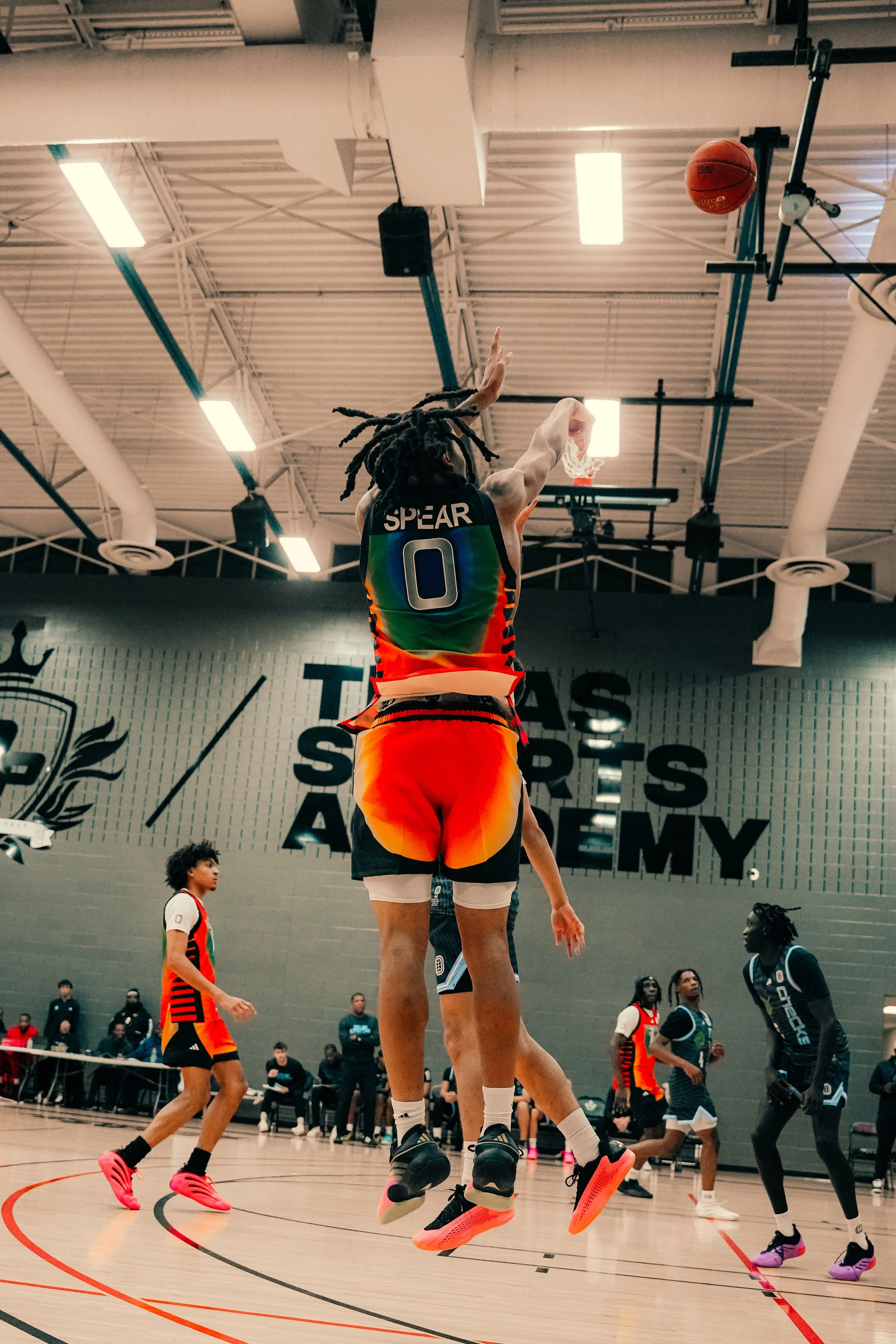 Basketball game with players jumping to catch the ball, multiple players on the court, spectators and coaches in the background, inside a gym with high ceiling and lights.