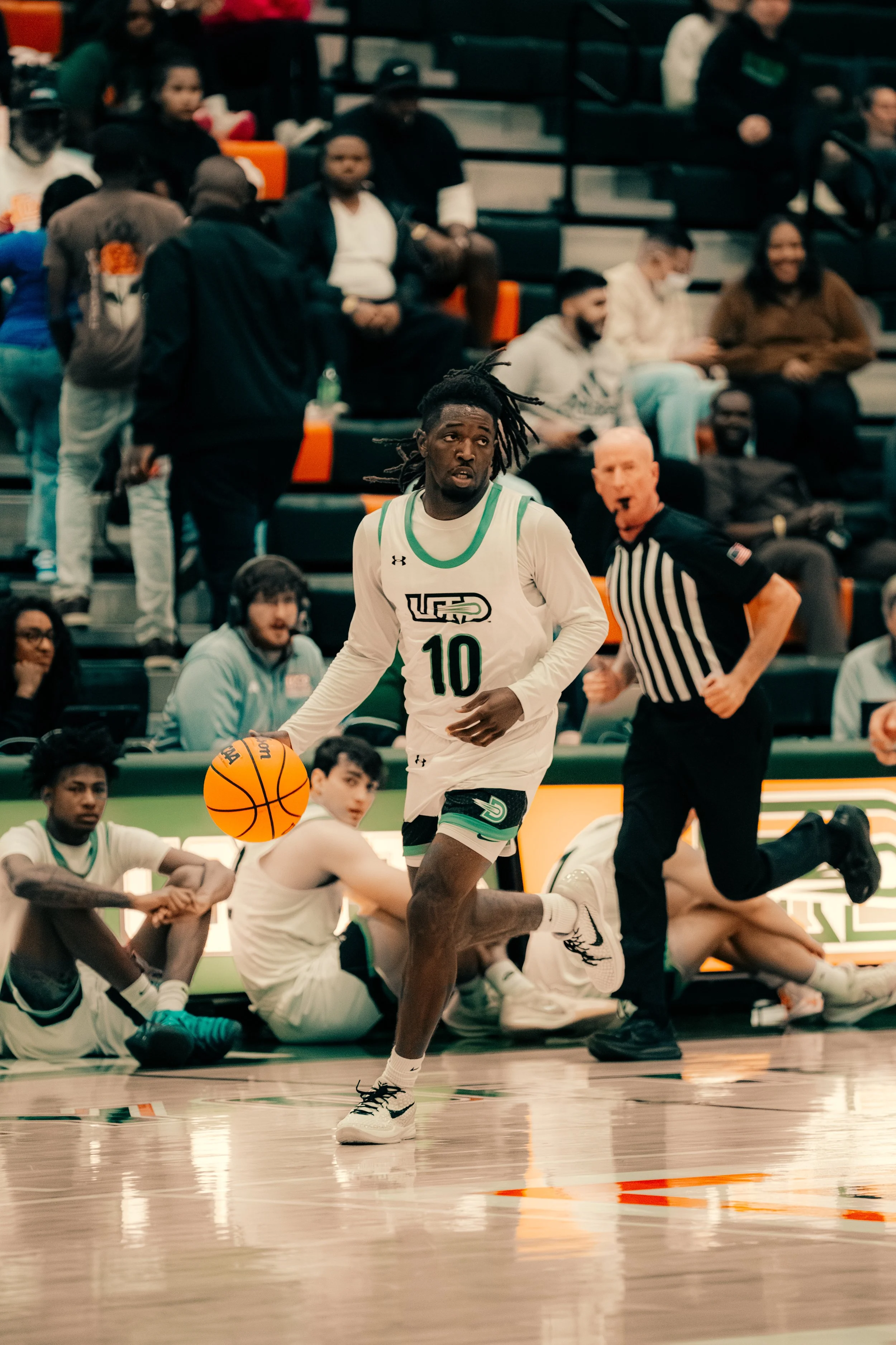 A basketball player wearing a white jersey with the number 10 dribbling the ball on the court during a game. In the background, there are spectators and team members sitting on the bench, with some sitting on the floor and others in the stands, watch