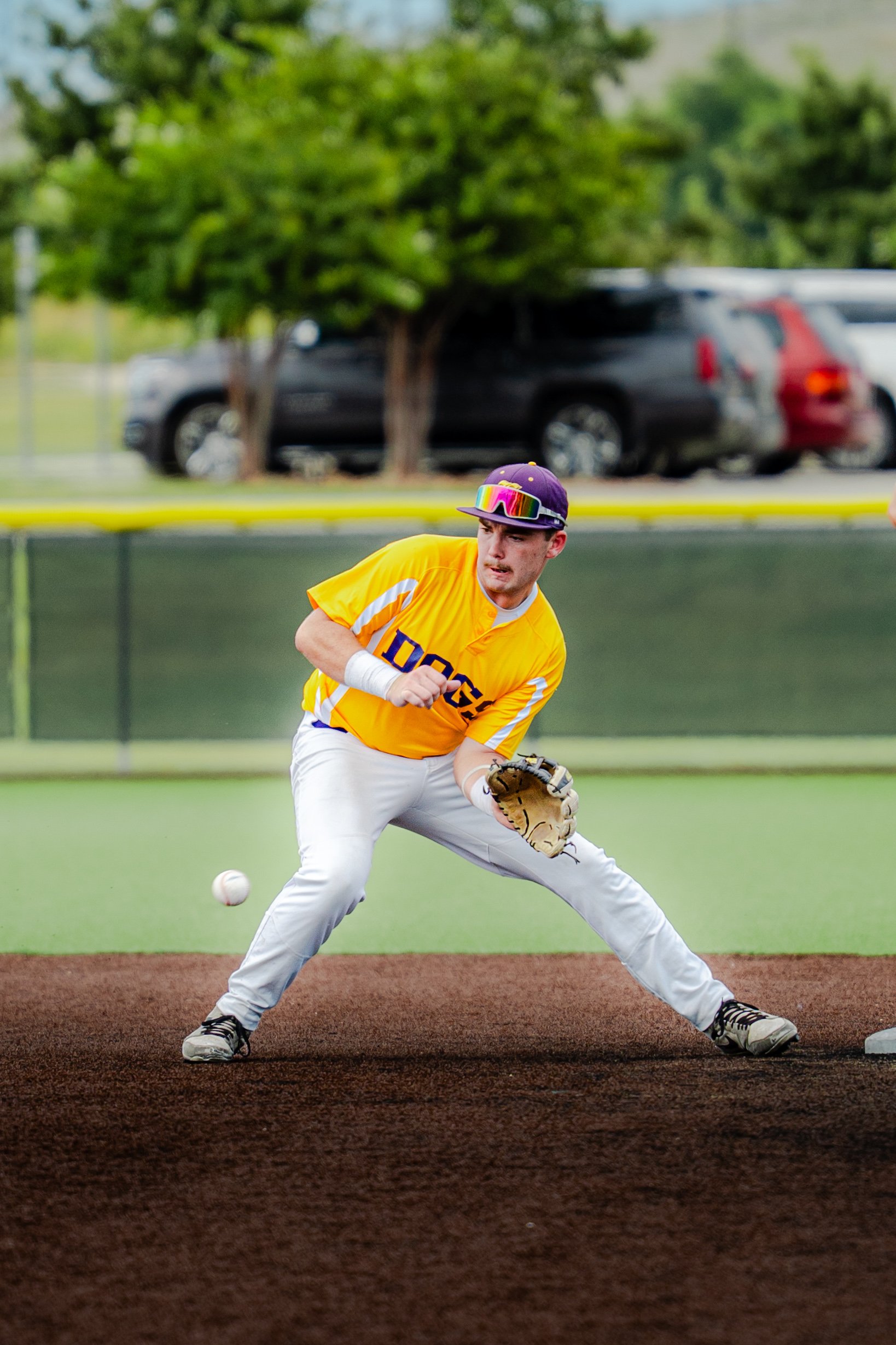A baseball player in a yellow jersey and white pants is fielding a ground ball on a baseball field, with a green fence and parked cars in the background.
