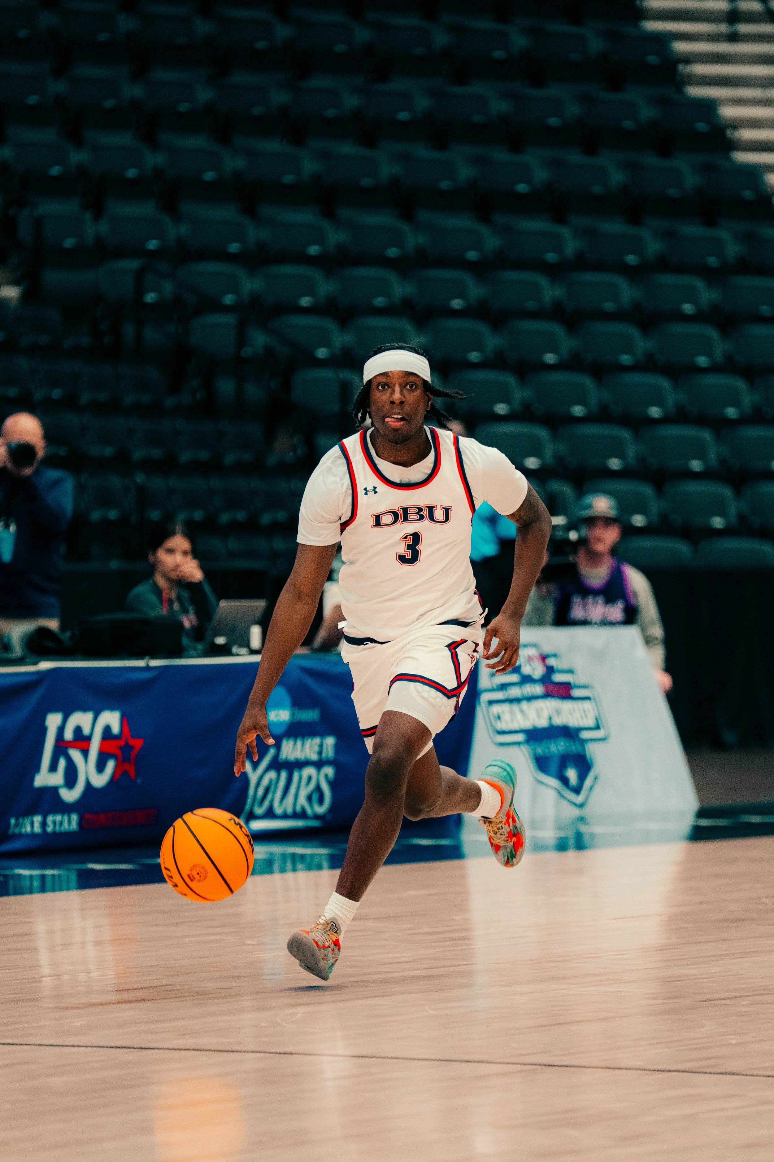 A male basketball player wearing a white jersey with 'DBU' and the number 3, running on the court during a game, dribbling an orange basketball, wearing colorful athletic shoes.
