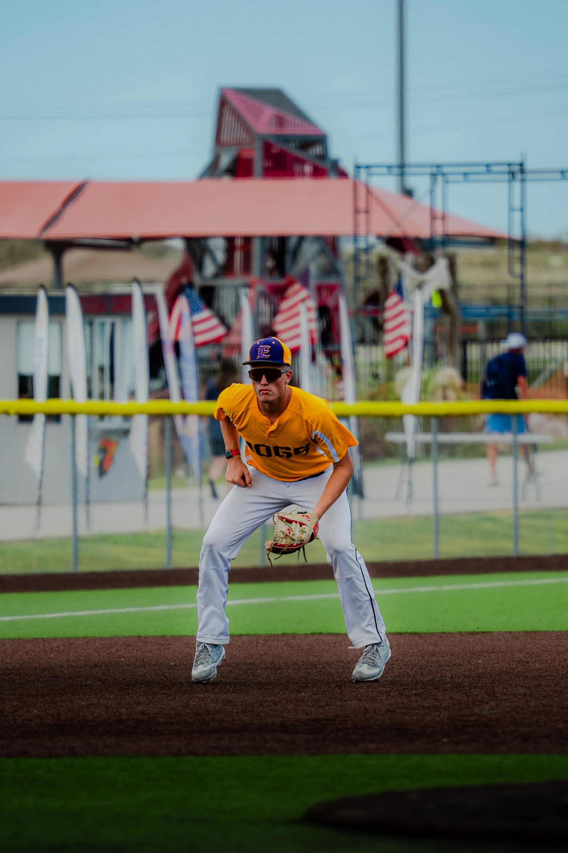 A baseball player wearing a yellow jersey, gray pants, sunglasses, and a blue cap with a yellow logo is positioned on the field, preparing to catch a ball. In the background, there is a red and pink amusement park ride with American flags, and some p