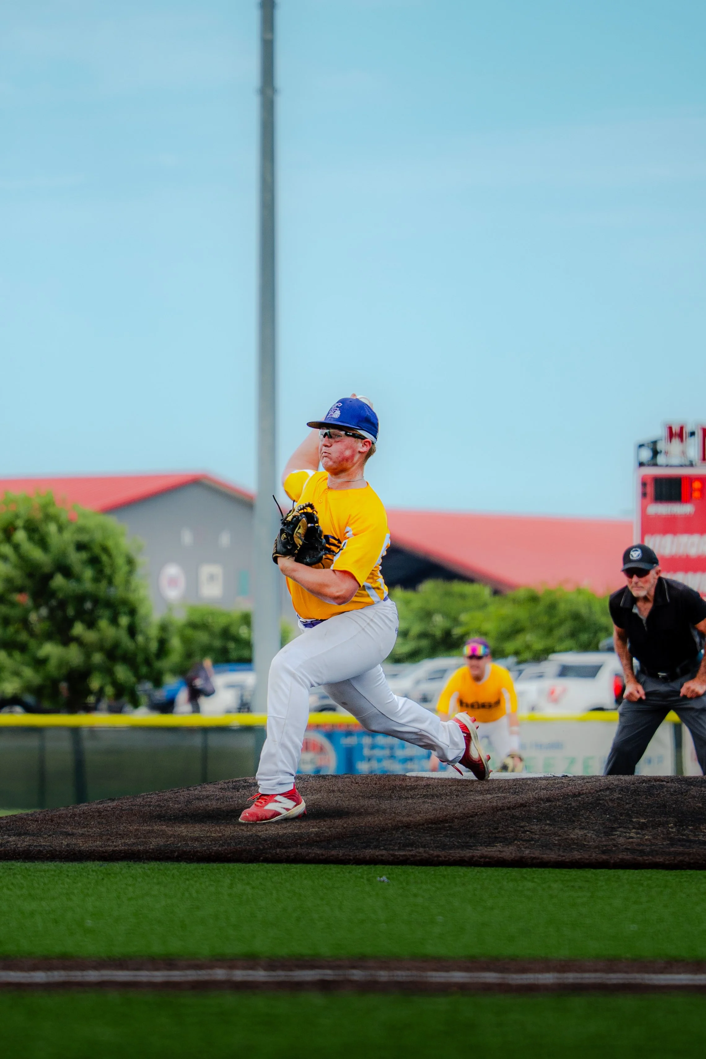 A baseball player in a yellow jersey and white pants is pitching on the mound, with other players and a coach in the background at a baseball field.