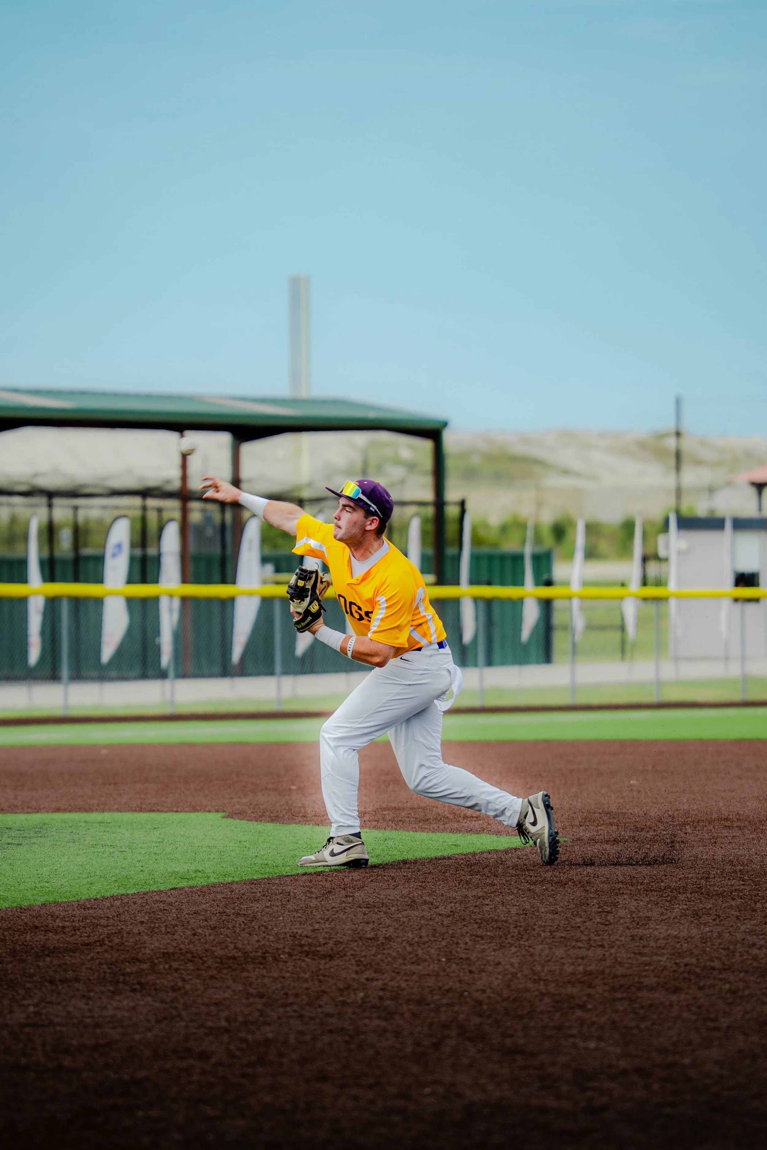 A baseball player in a yellow jersey and white pants is preparing to catch a ball on a baseball field, with a blue sky and some structures in the background.