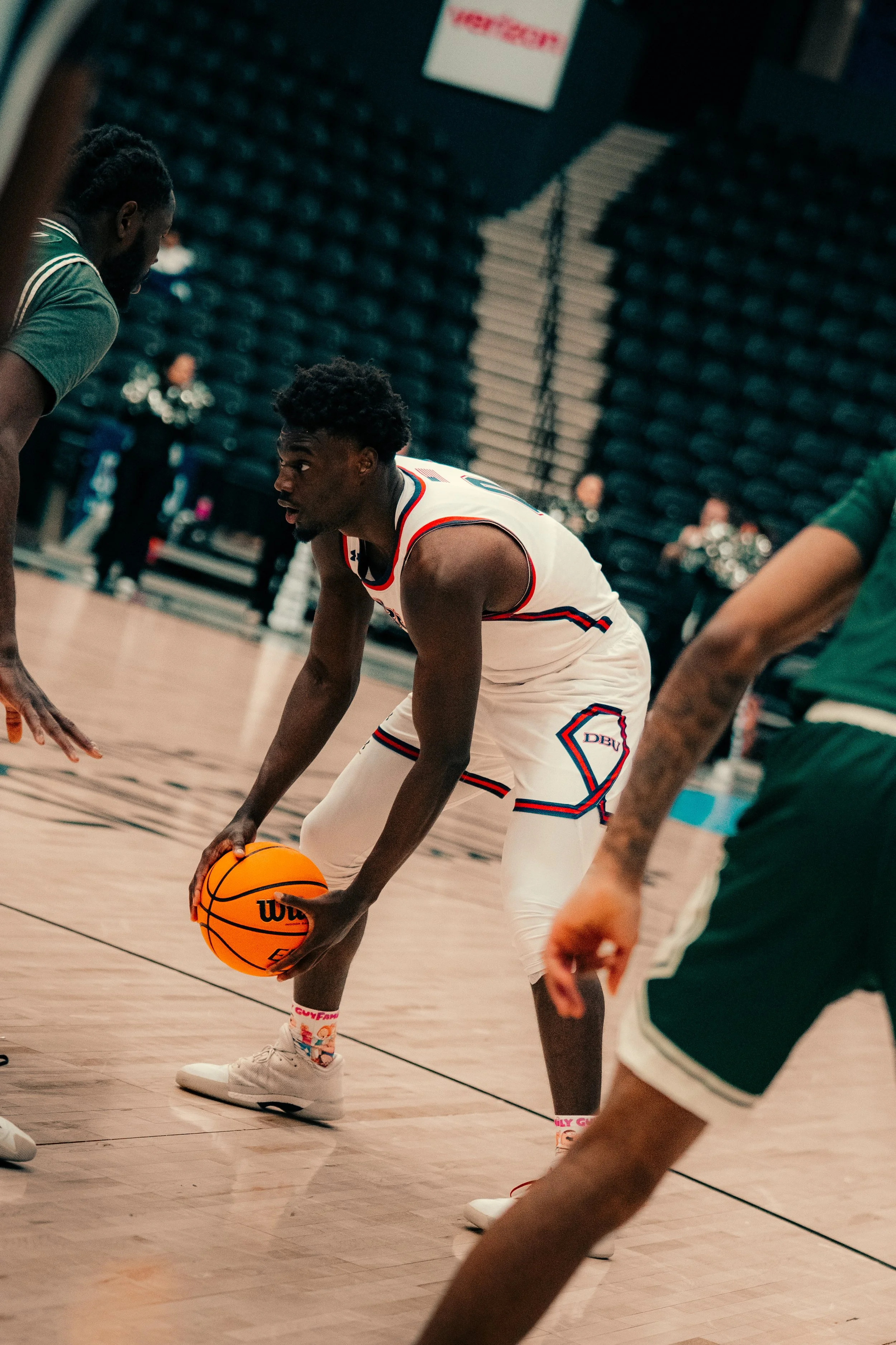 A basketball player in a white jersey with blue and red details is bending down to pick up an orange basketball on a basketball court. Other players and spectators are visible in the background.