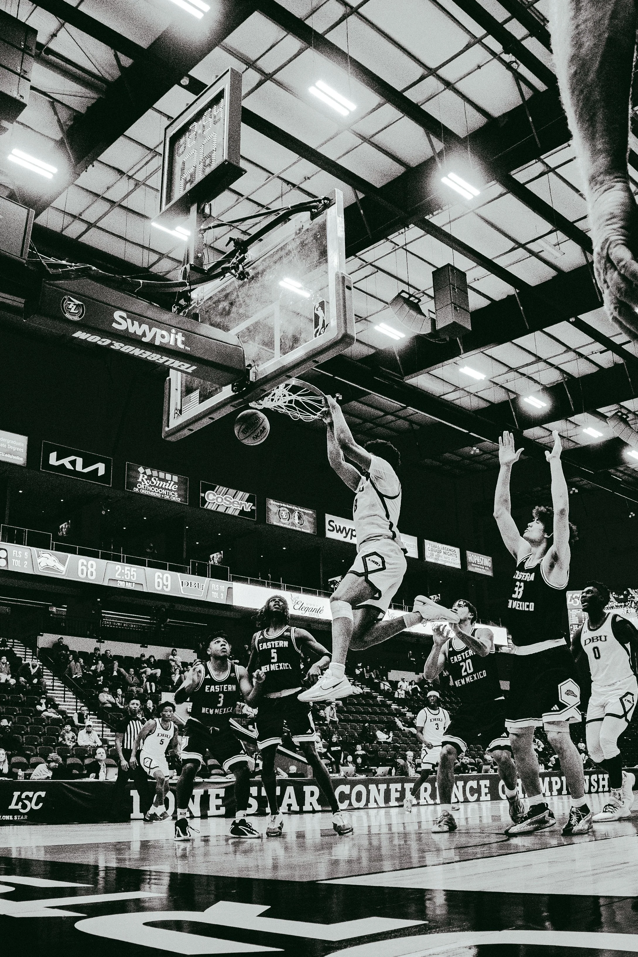Basketball game in progress with players jumping for the ball under the hoop, spectators in the background, and scoreboard showing scores and time.