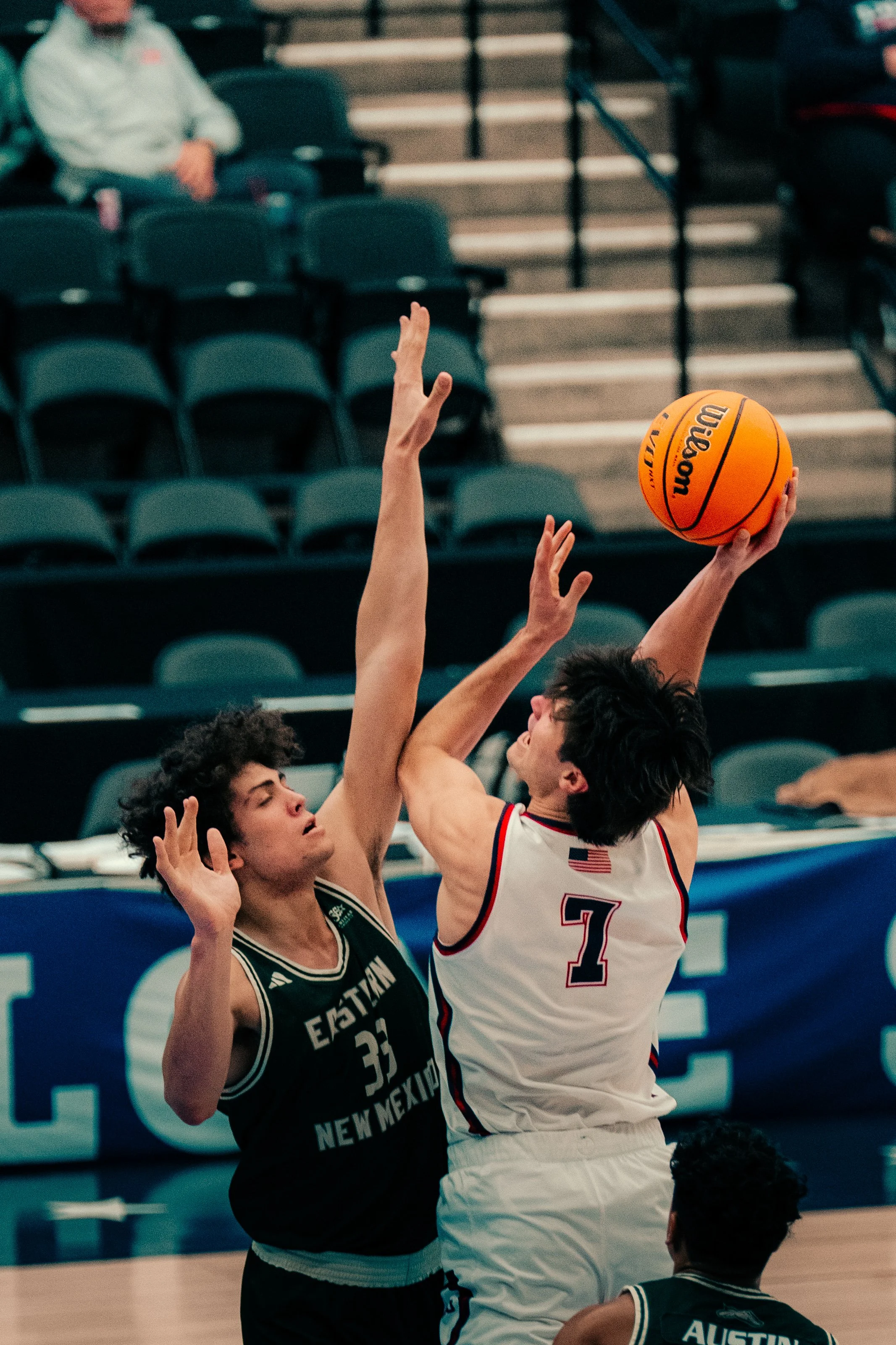 Two basketball players from opposing teams compete for the ball during a game. One player is wearing a black jersey with white lettering that reads "EASTERN NEW MEXICO" and the number 33. The other player is wearing a white jersey with red and blue a