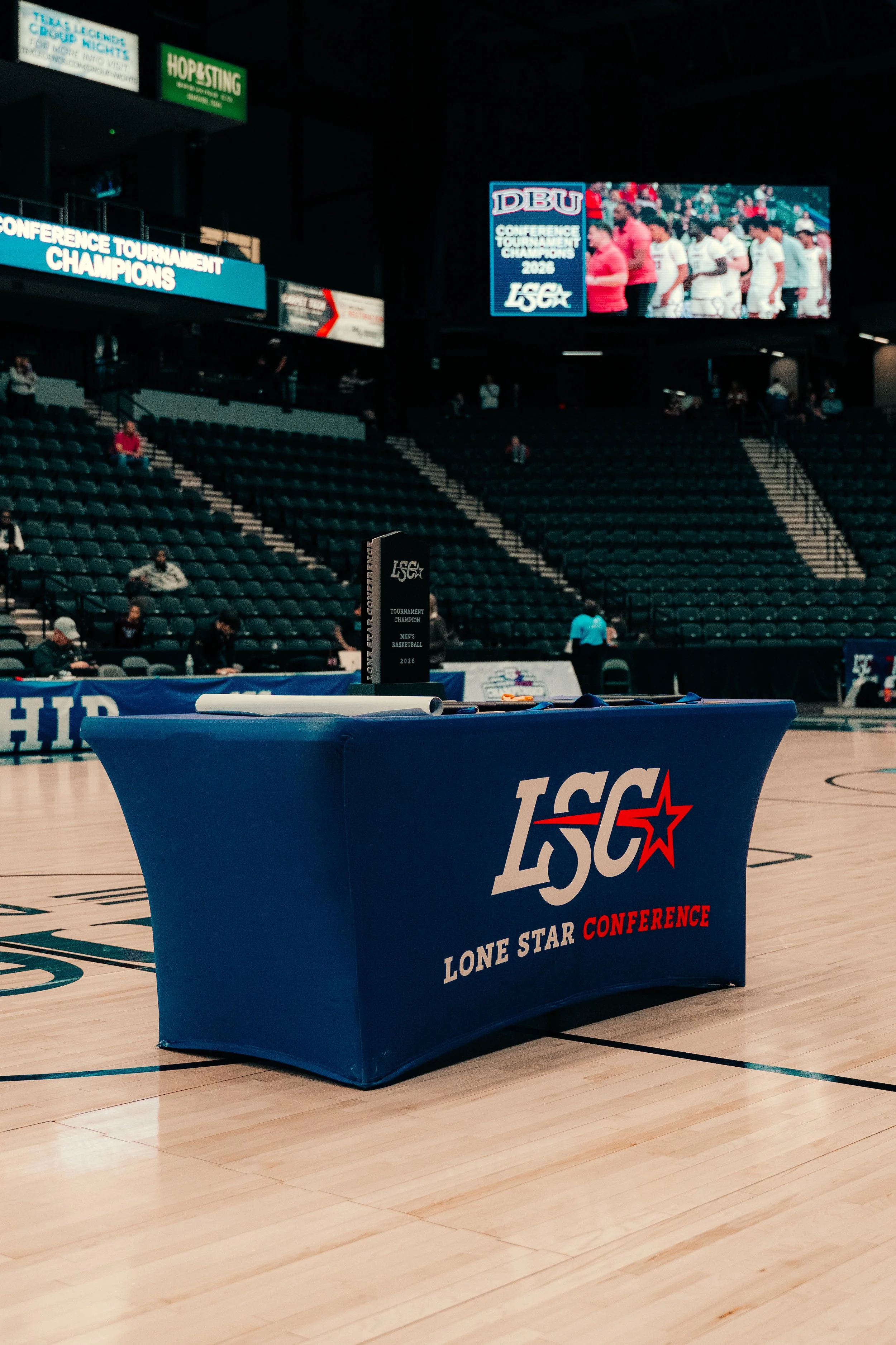 Indoor basketball arena with a blue table cover displaying 'Lone Star Conference' and the league's logo, a black trophy on top, and a large LED screen in the background showing a lineup of basketball players. Some spectators are seated in the stands.