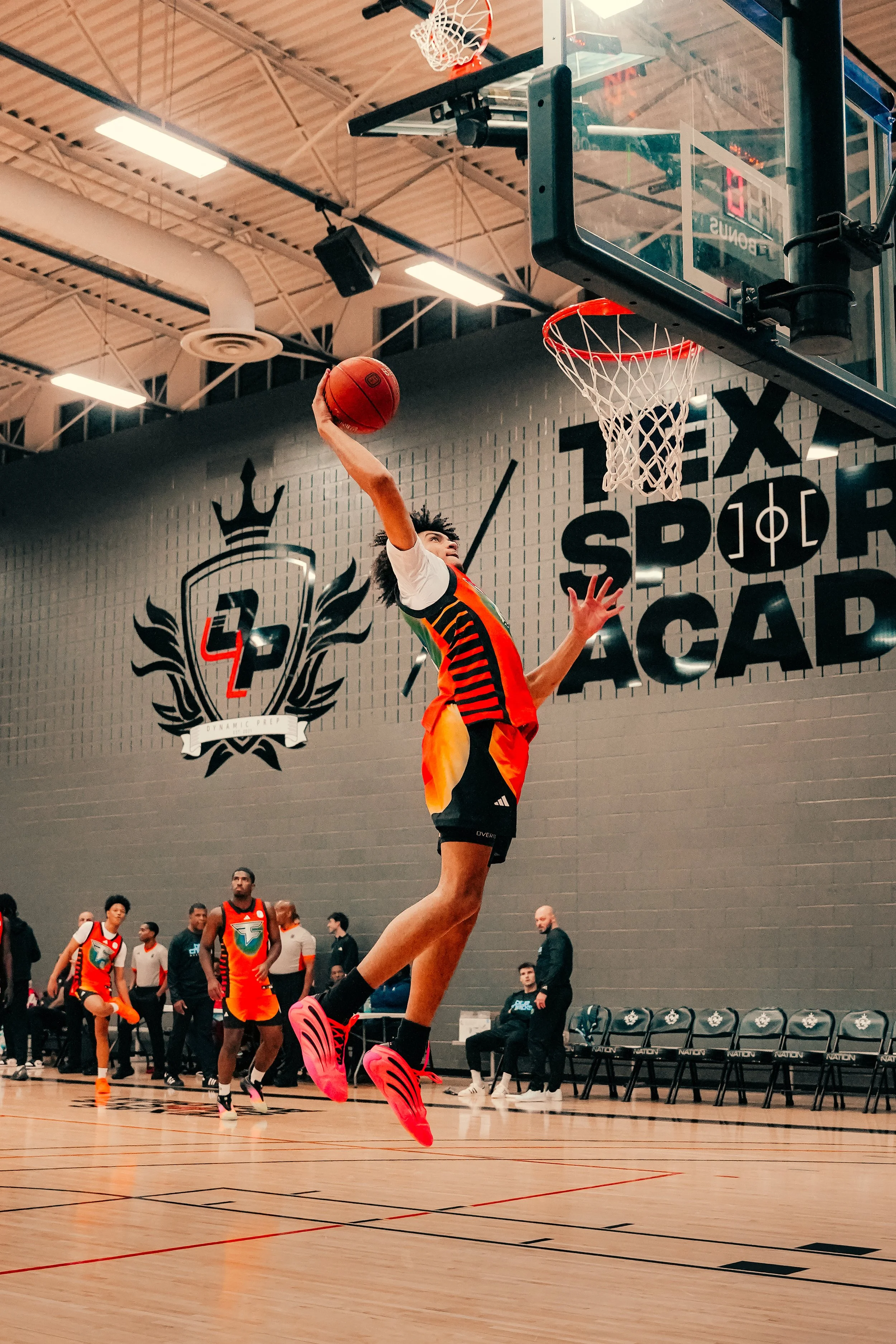 A basketball player in mid-air about to make a dunk at an indoor basketball court with other players and spectators in the background.