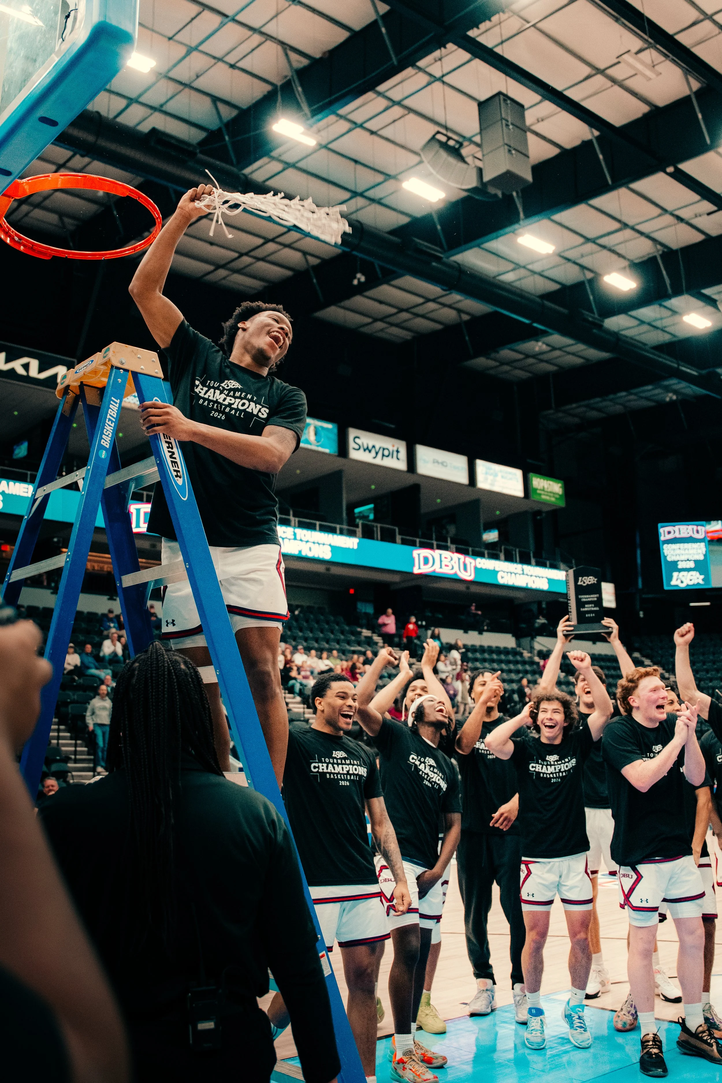 Group of basketball players celebrating on the court after a game, some smiling, wearing black shirts and white shorts, with one player standing on a ladder.