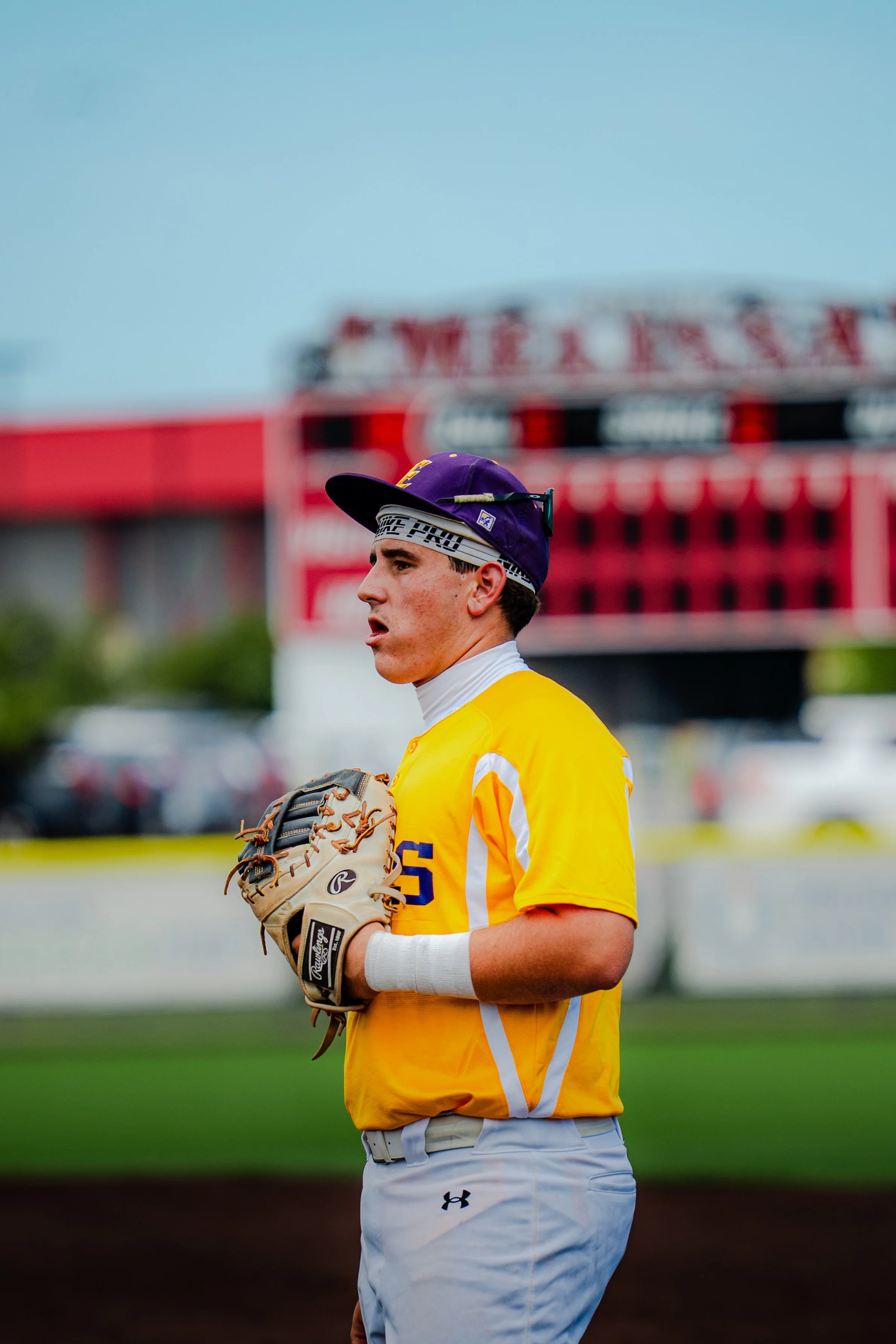 A young baseball player in a yellow jersey and white Baseball pants standing on the field with a baseball glove, wearing a purple cap and white headband, with a blurred stadium background.
