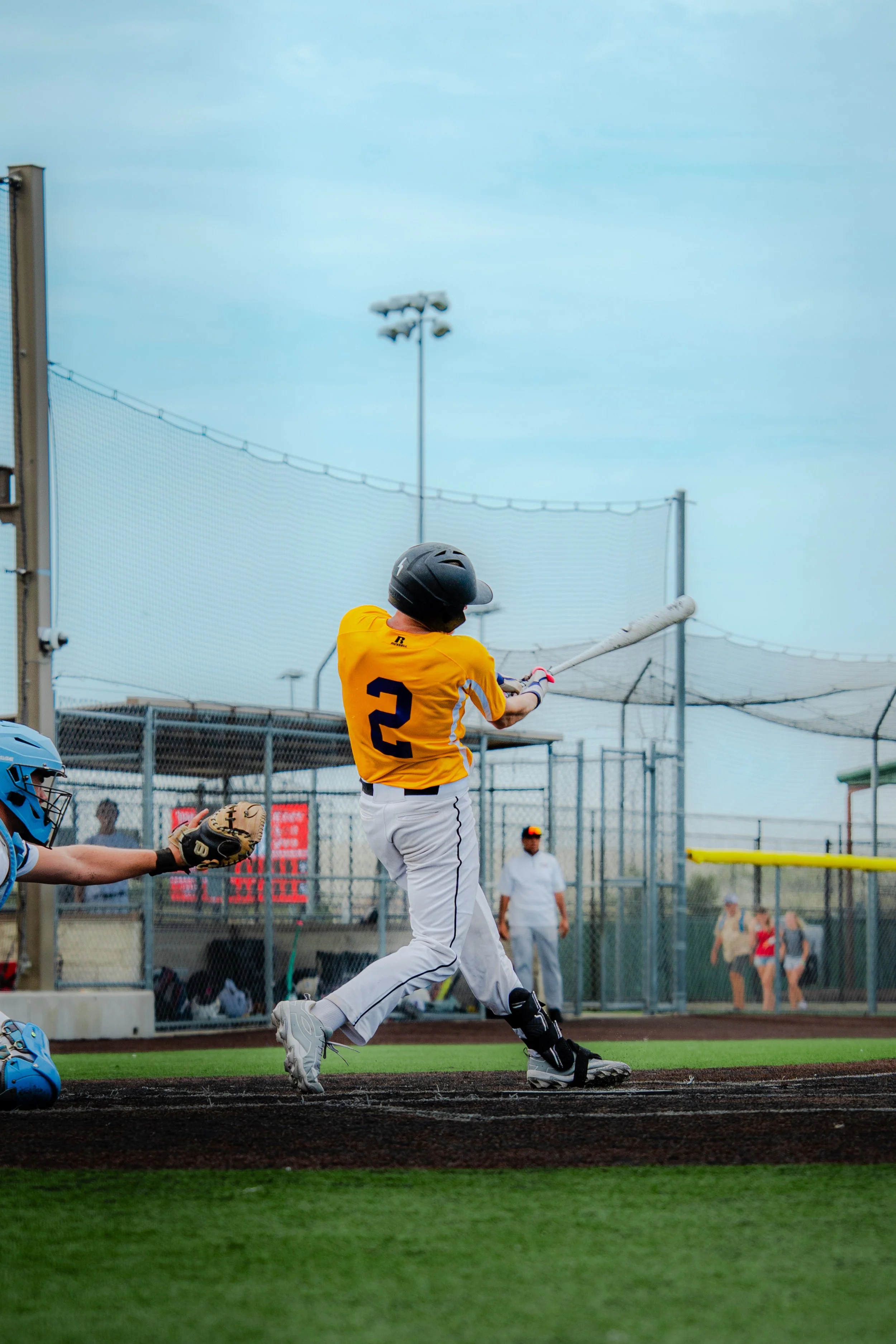 A baseball player in a yellow jersey with the number 2 swings a bat on a baseball field. A catcher in blue gear is crouched behind home plate trying to catch the ball. A person dressed in white stands in the background near the fence.