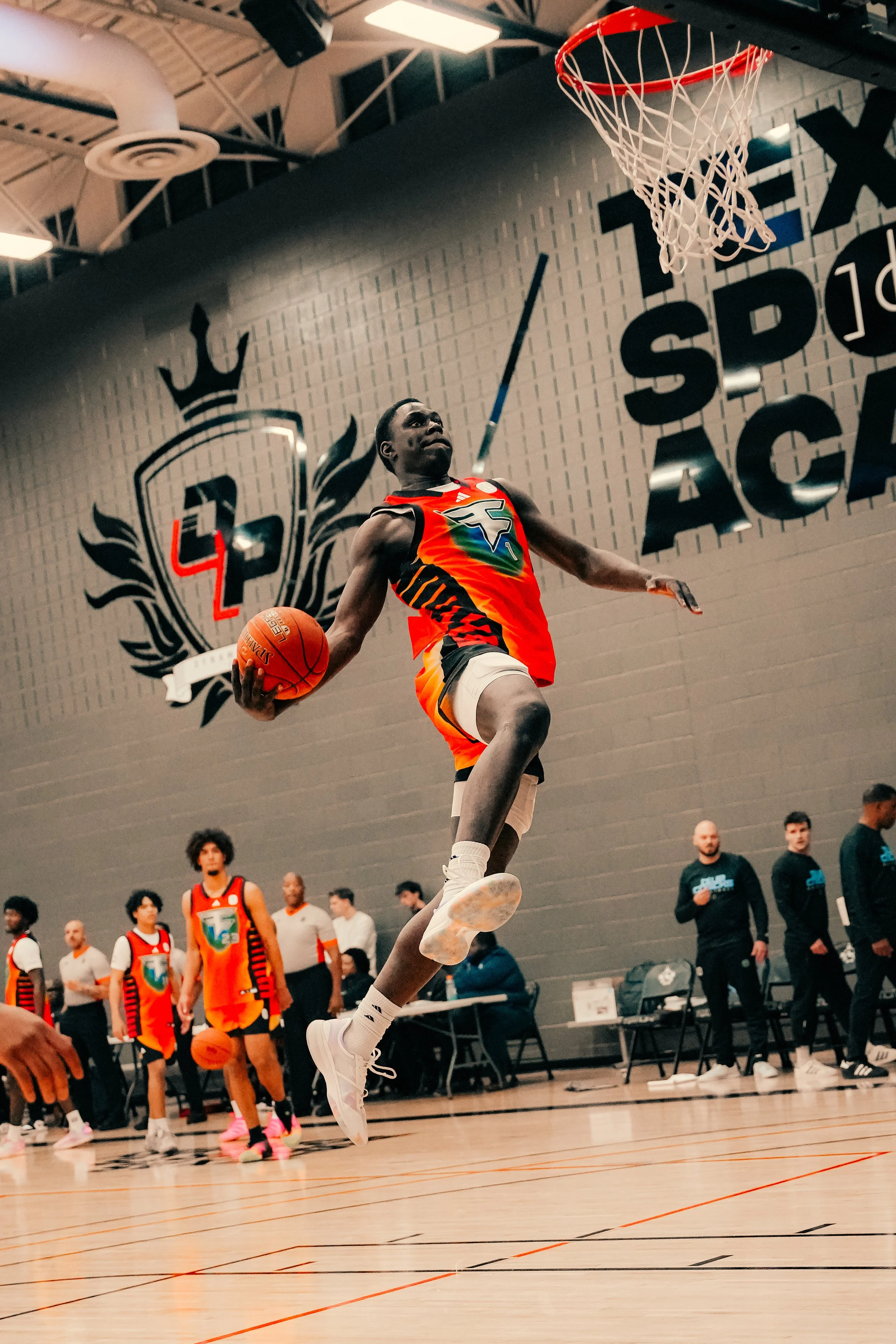 A basketball player in a colorful uniform jumps towards the hoop during a game, with spectators and other players watching in the background inside a gymnasium.