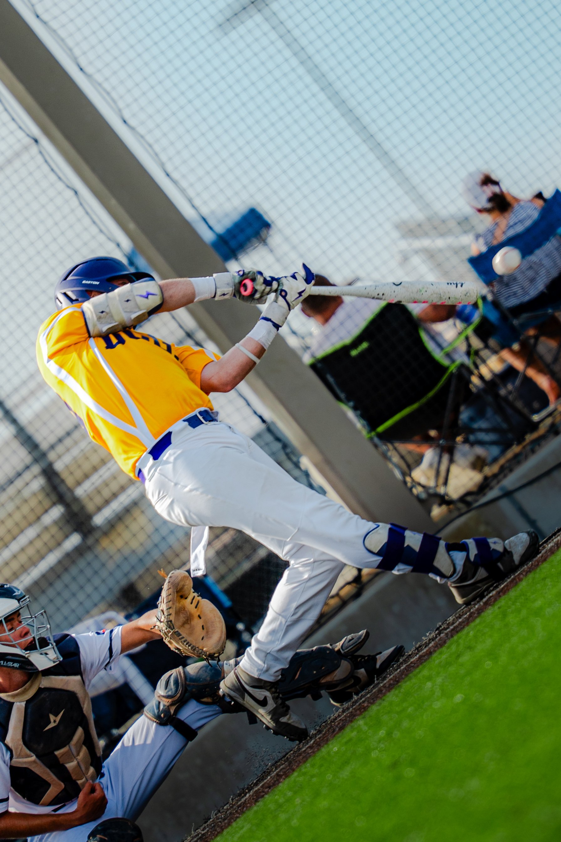 A baseball player in a yellow jersey, white pants, and a helmet swings a bat at a pitched ball during a game. The catcher in a protective suit, mask, and mitt is crouched behind him, ready to catch the ball. The game is played on a field with green t