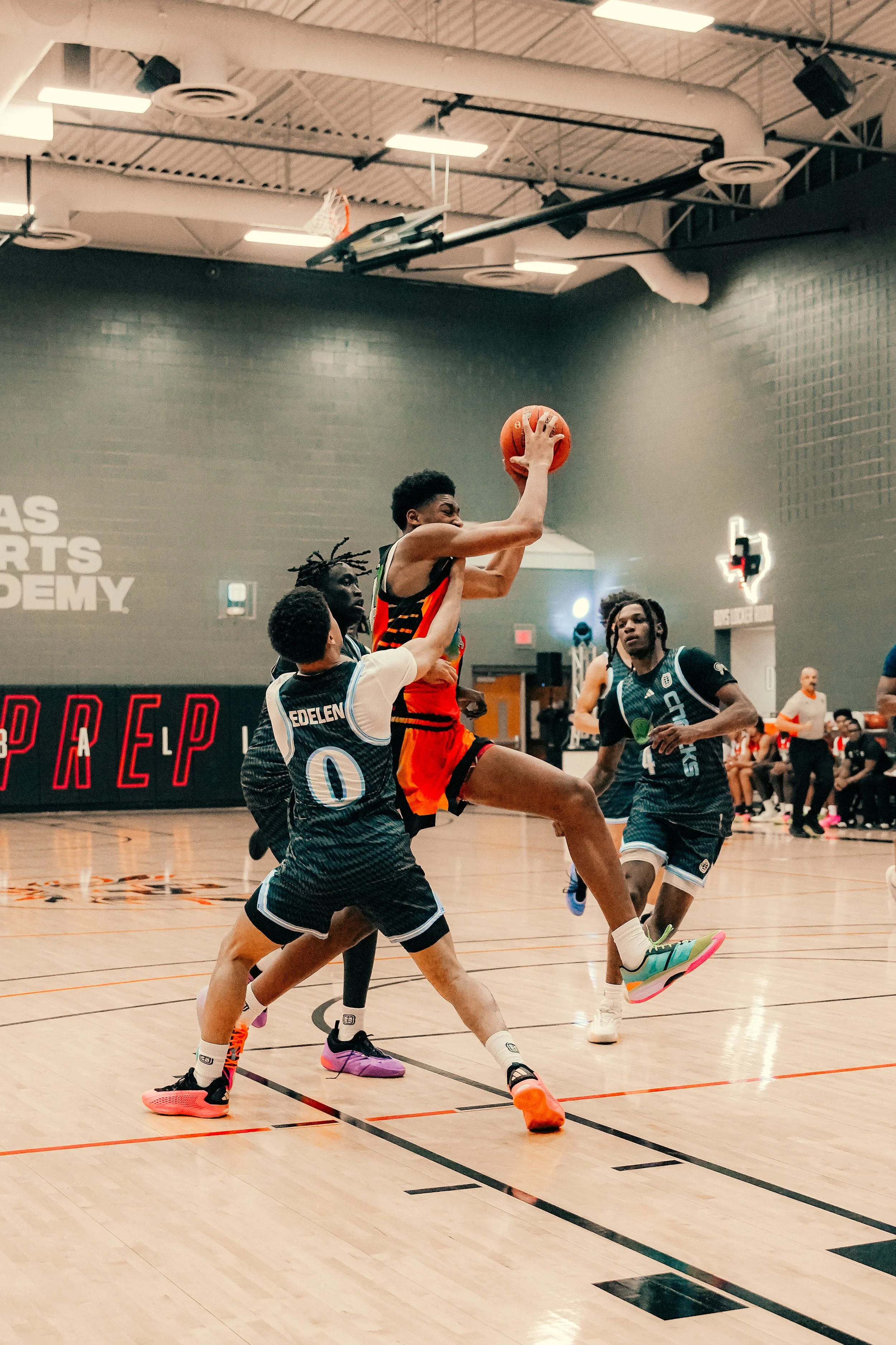 Two basketball players are jumping to grab the ball during a game in an indoor gym. One player is wearing orange and black, while the other is in a black uniform with white and blue accents. Several other players watch on, and a referee or coach stan