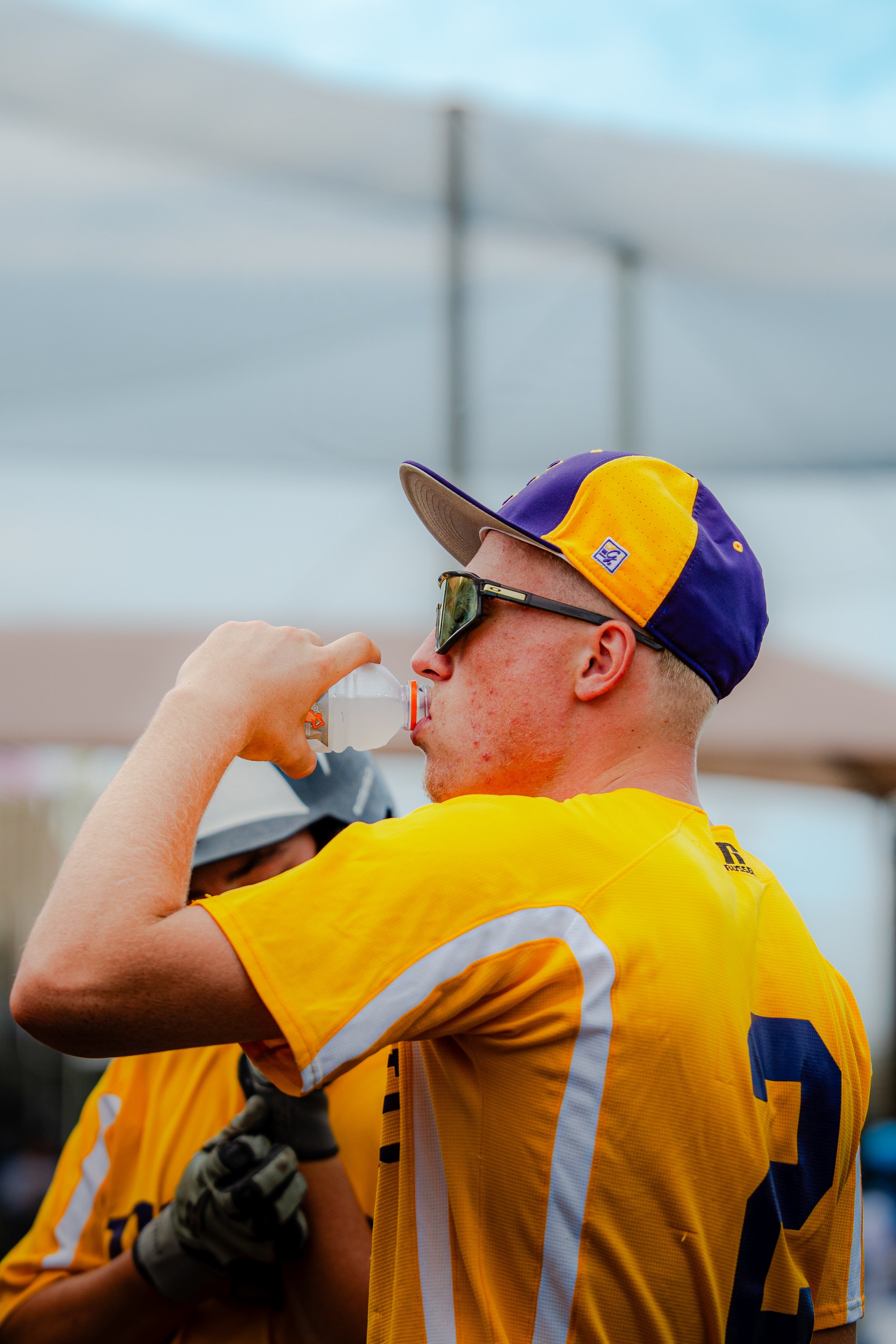 A person wearing a yellow sports jersey, sunglasses, and a purple, yellow, and grey cap, drinking from a plastic water bottle. Another person with a helmet and gloves is partly visible.