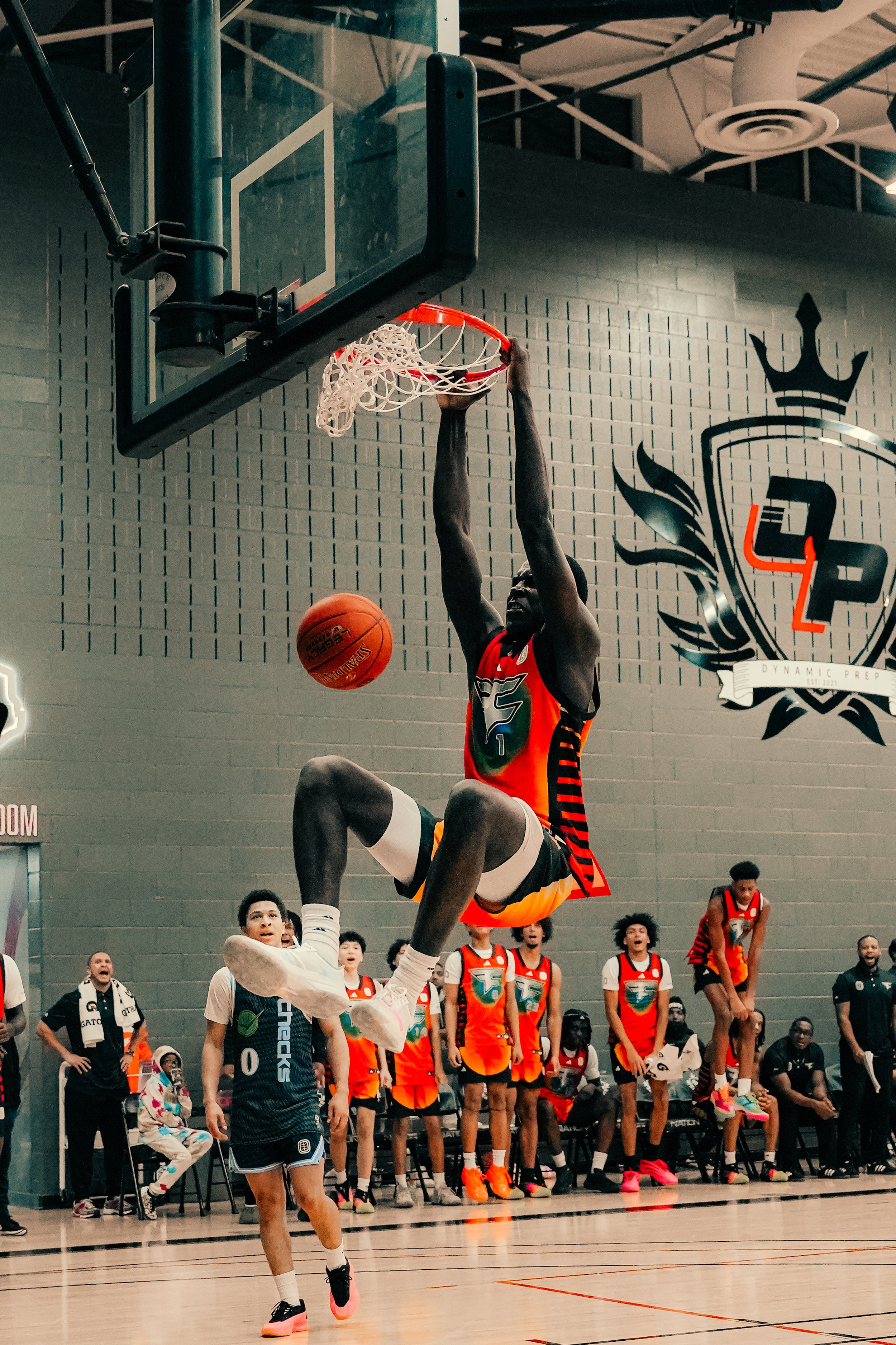 A basketball player in a red and black uniform is dunking the ball into the hoop, while another player in a blue uniform attempts to block the shot. Several people in sportswear and jerseys are watching from the sidelines and background inside a gymn
