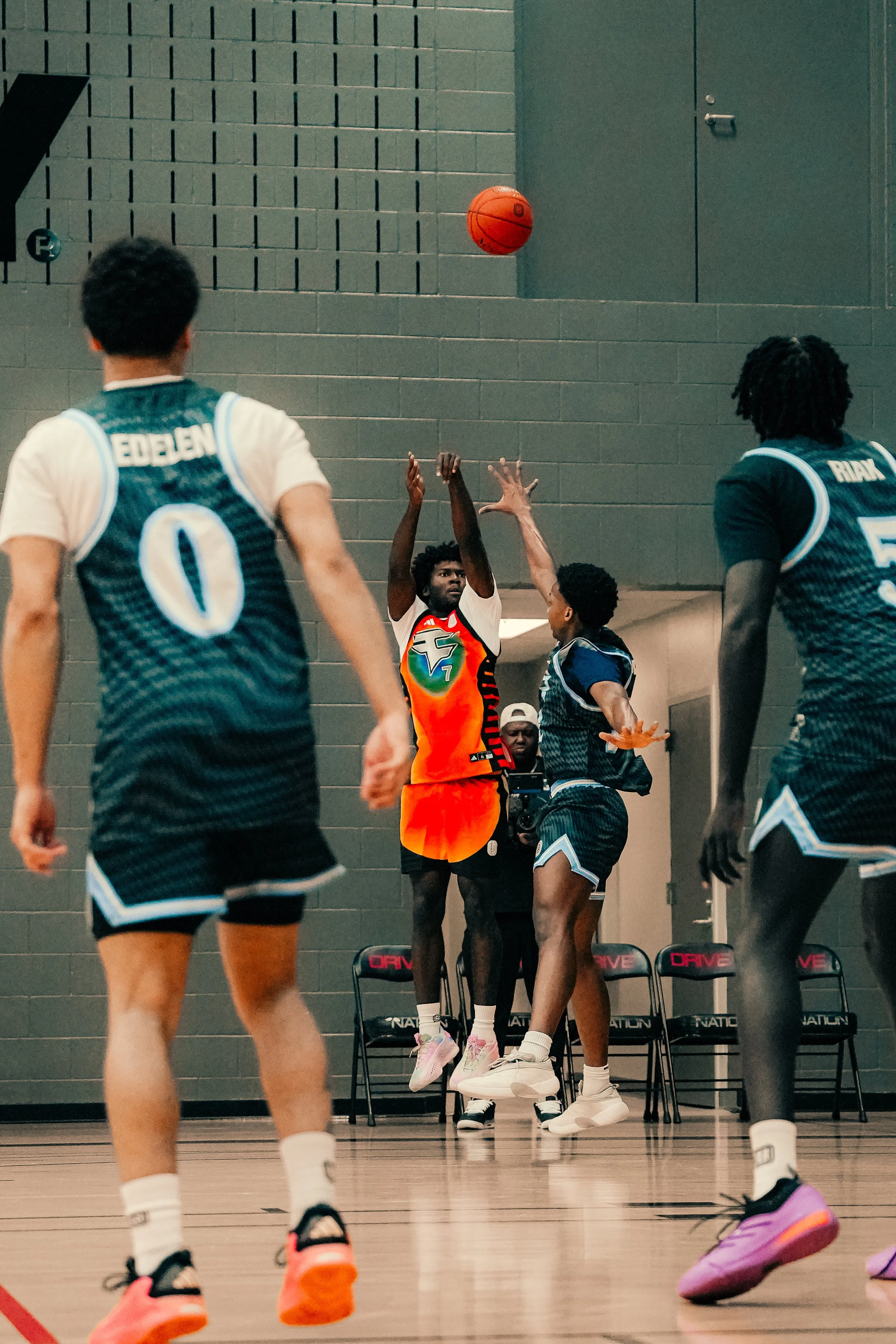 A basketball game is happening indoors with players jumping for the ball. Two players are jumping to reach the ball, one wearing an orange jersey, and the others are in dark jerseys. Spectators are visible on chairs in the background.