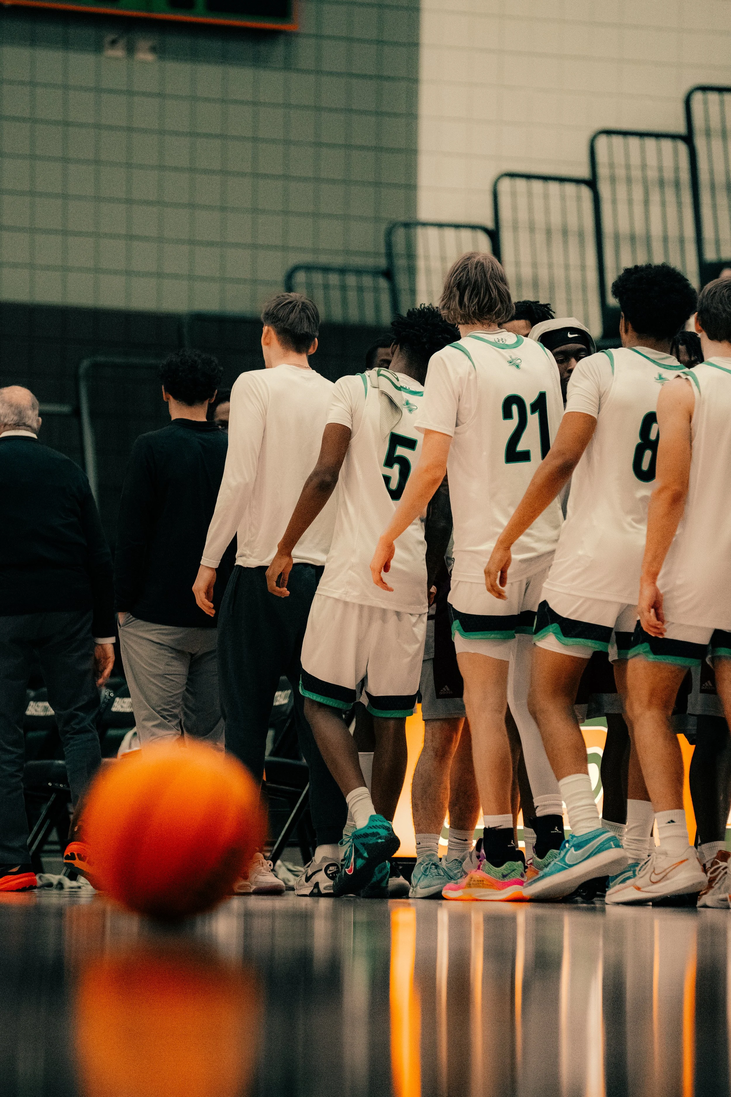 A group of basketball players wearing white jerseys with green accents are huddled together on an indoor court, with a basketball on the ground in the foreground and various people and equipment visible in the background.