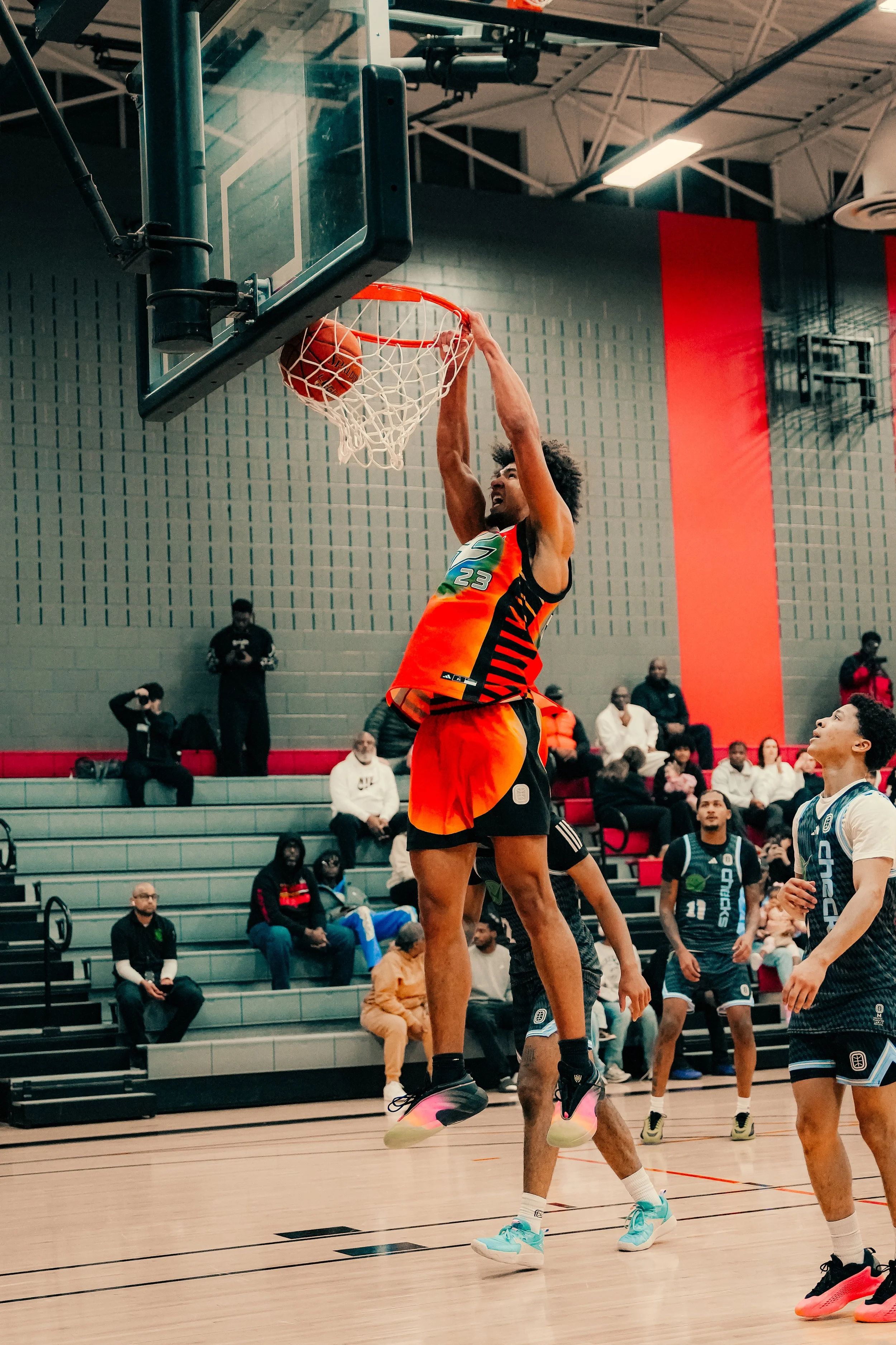 A basketball player in orange and black uniform, wearing number 23, is jumping to shoot the basketball into the hoop during a game. The scene takes place in an indoor gym with spectators and other players watching.
