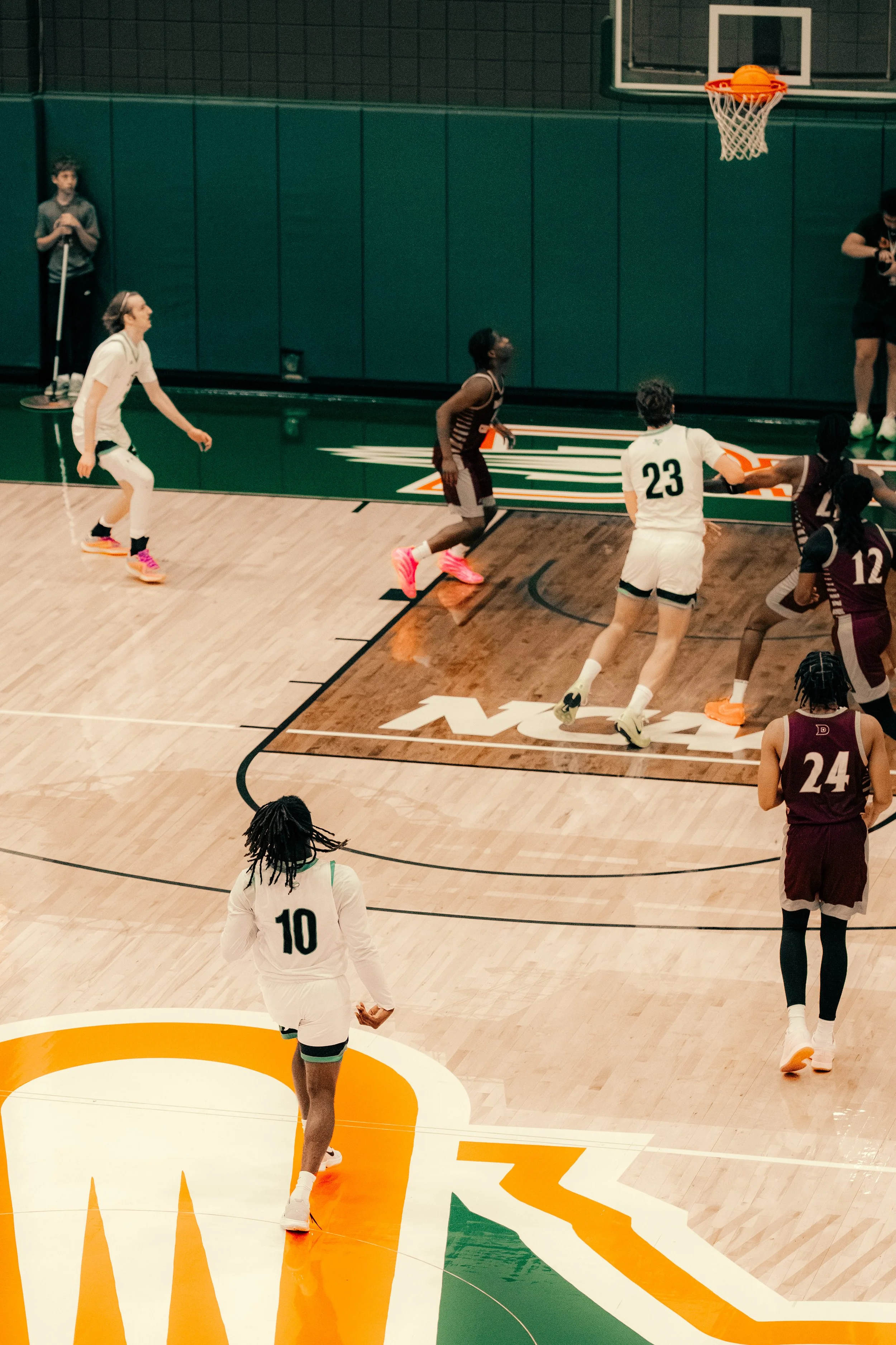 A group of basketball players on a court, with some preparing for a free throw and others standing nearby. The scene takes place in an indoor gym with a basketball hoop in the background.