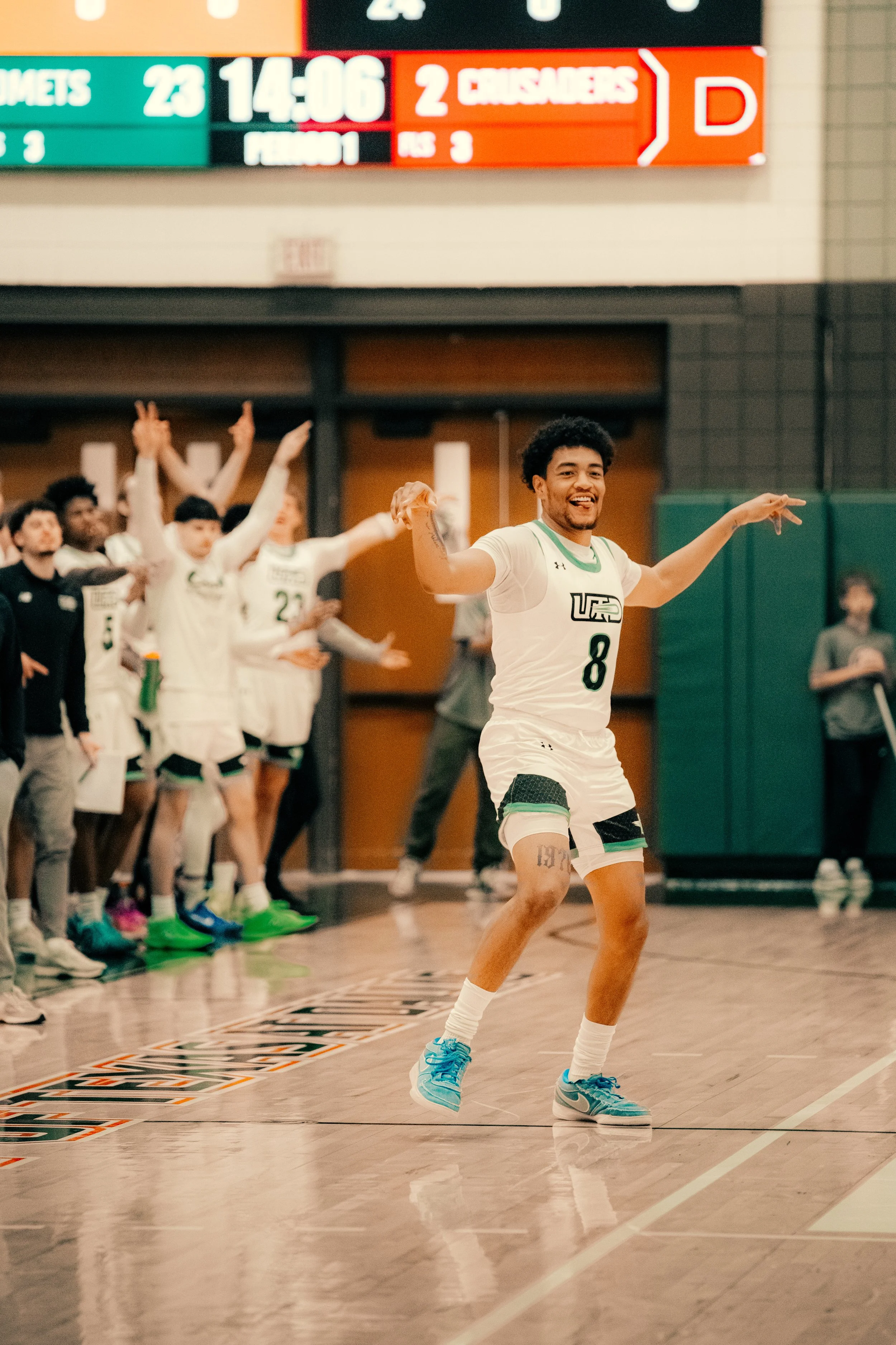 A volleyball player in a white uniform with the number 8 is celebrating on the court with arms outstretched, teammates in white uniforms with the team logo are cheering in the background, and a digital scoreboard is visible above.