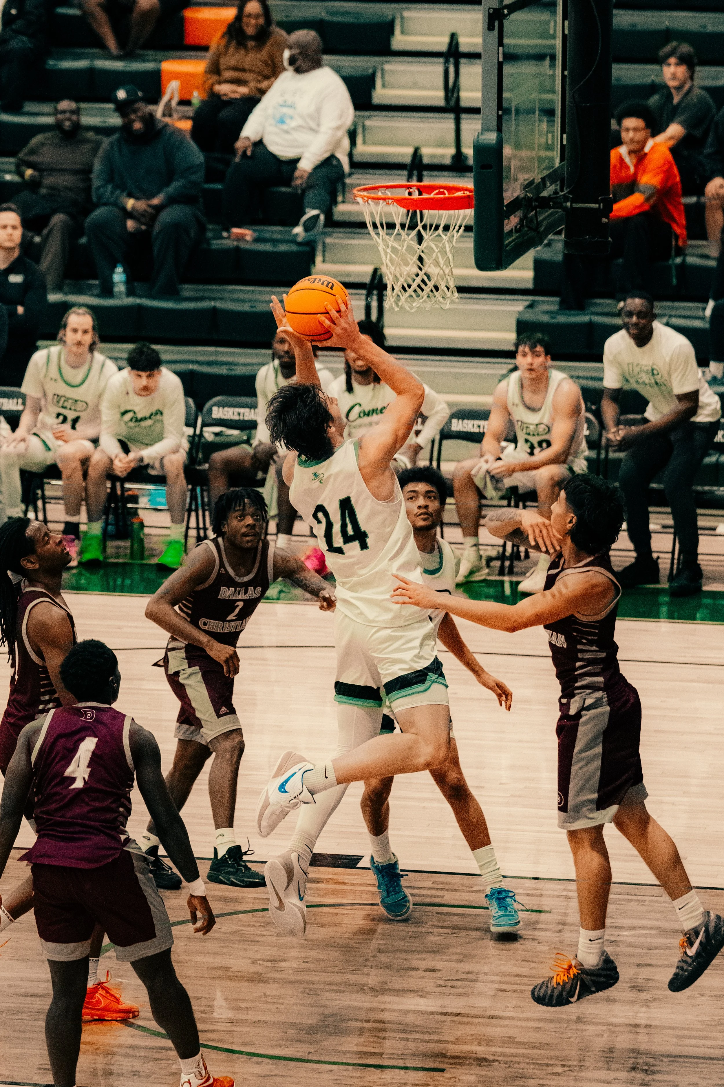 A basketball game in progress inside a gymnasium, with players in white and maroon jerseys and spectators seated in the background.