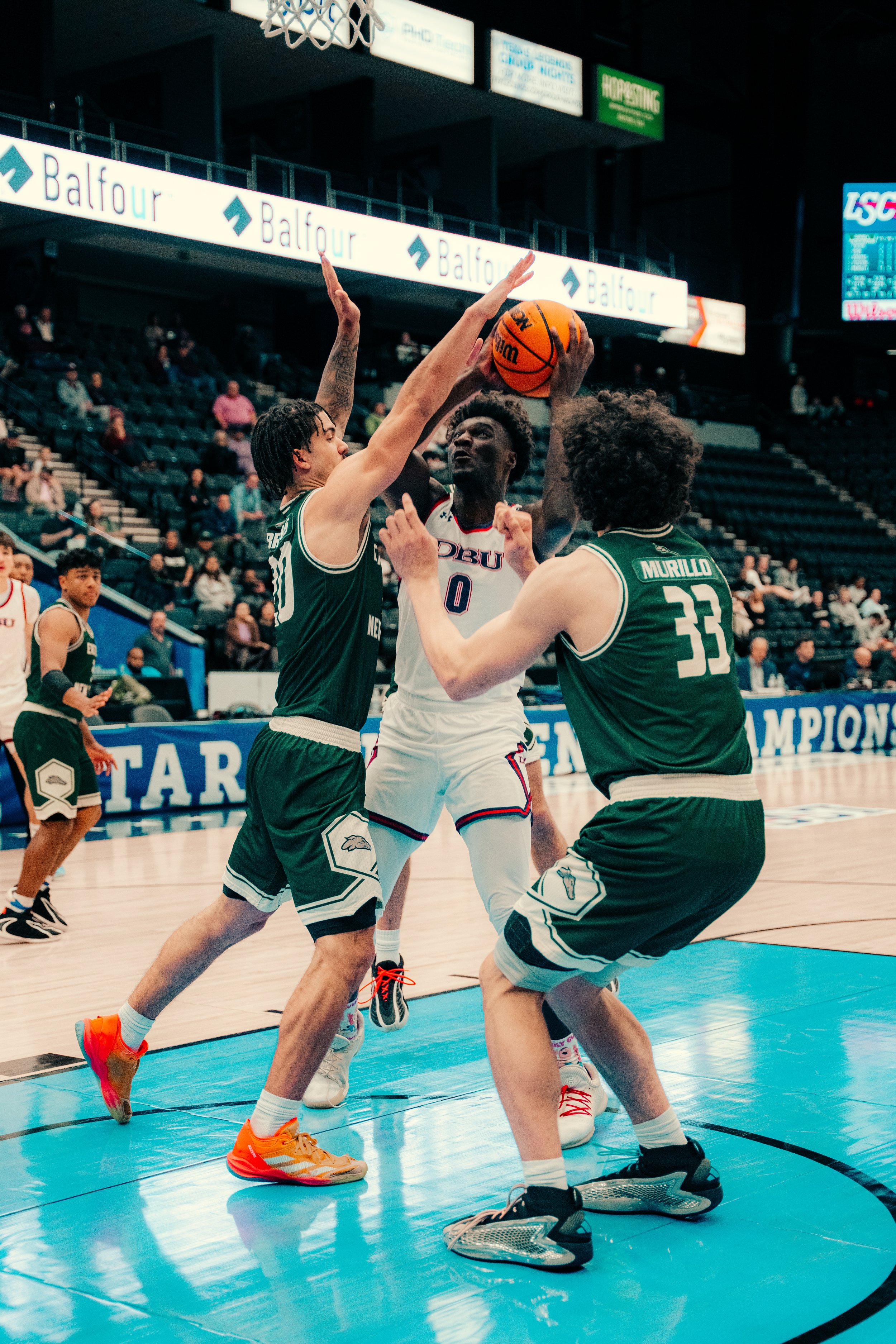 Two basketball players from opposing teams compete for possession of the ball near the basket, with spectators seated in the background.