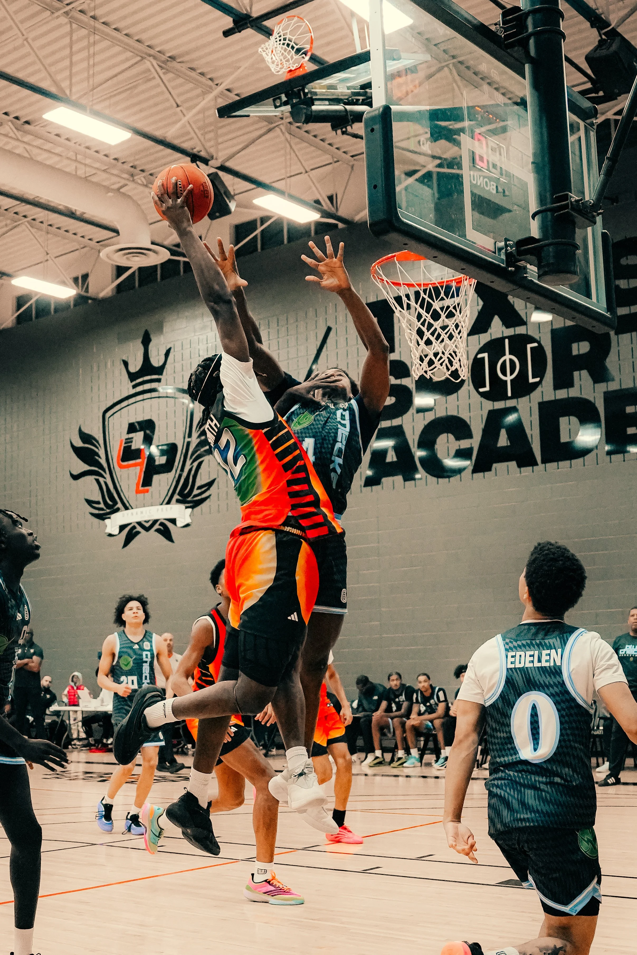 A basketball game in progress with players jumping for the ball near the hoop. The gym is filled with spectators and other players watching the action.