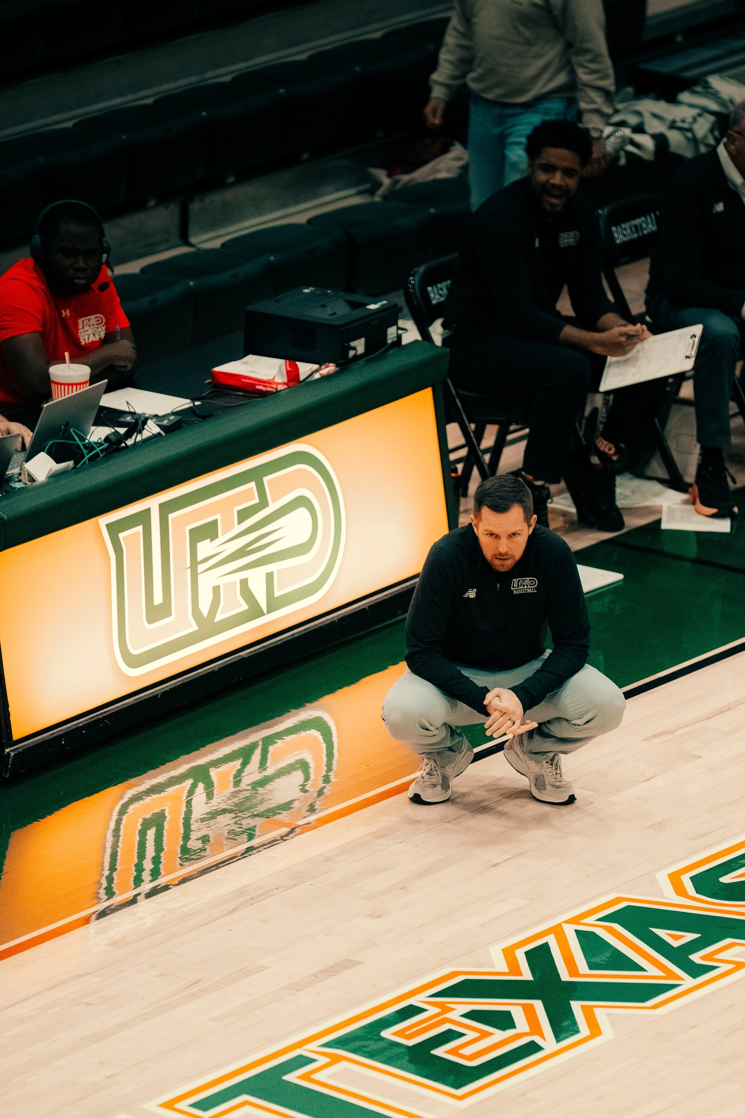 People sitting courtside at a basketball game, including a man crouching on the floor near the team logo, and others sitting behind the scorer's table with electronic equipment and drinks.