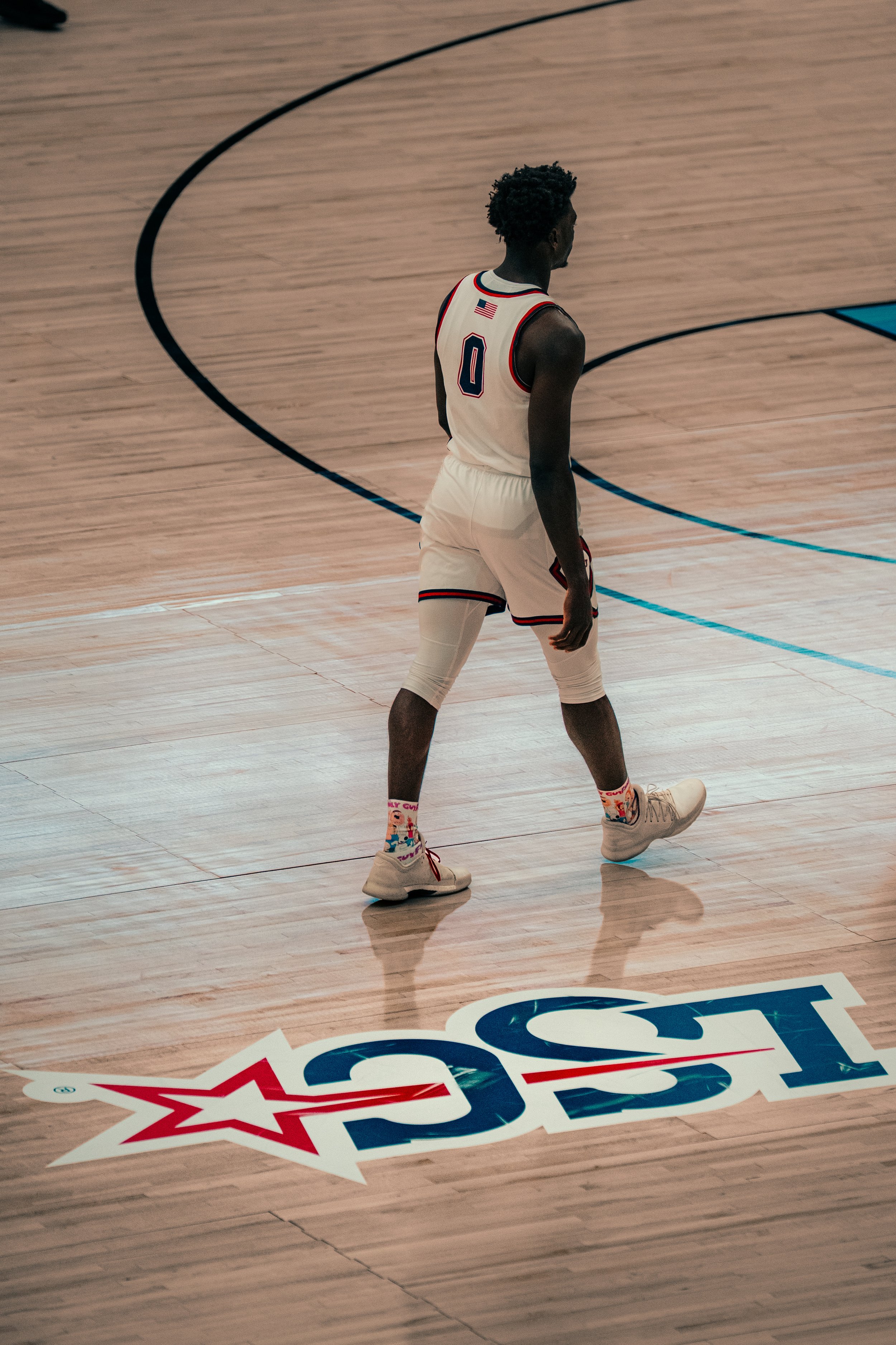 A basketball player on the court wearing a white jersey with the number 0, standing next to a large logo on the wooden floor that has a star and abstract design.