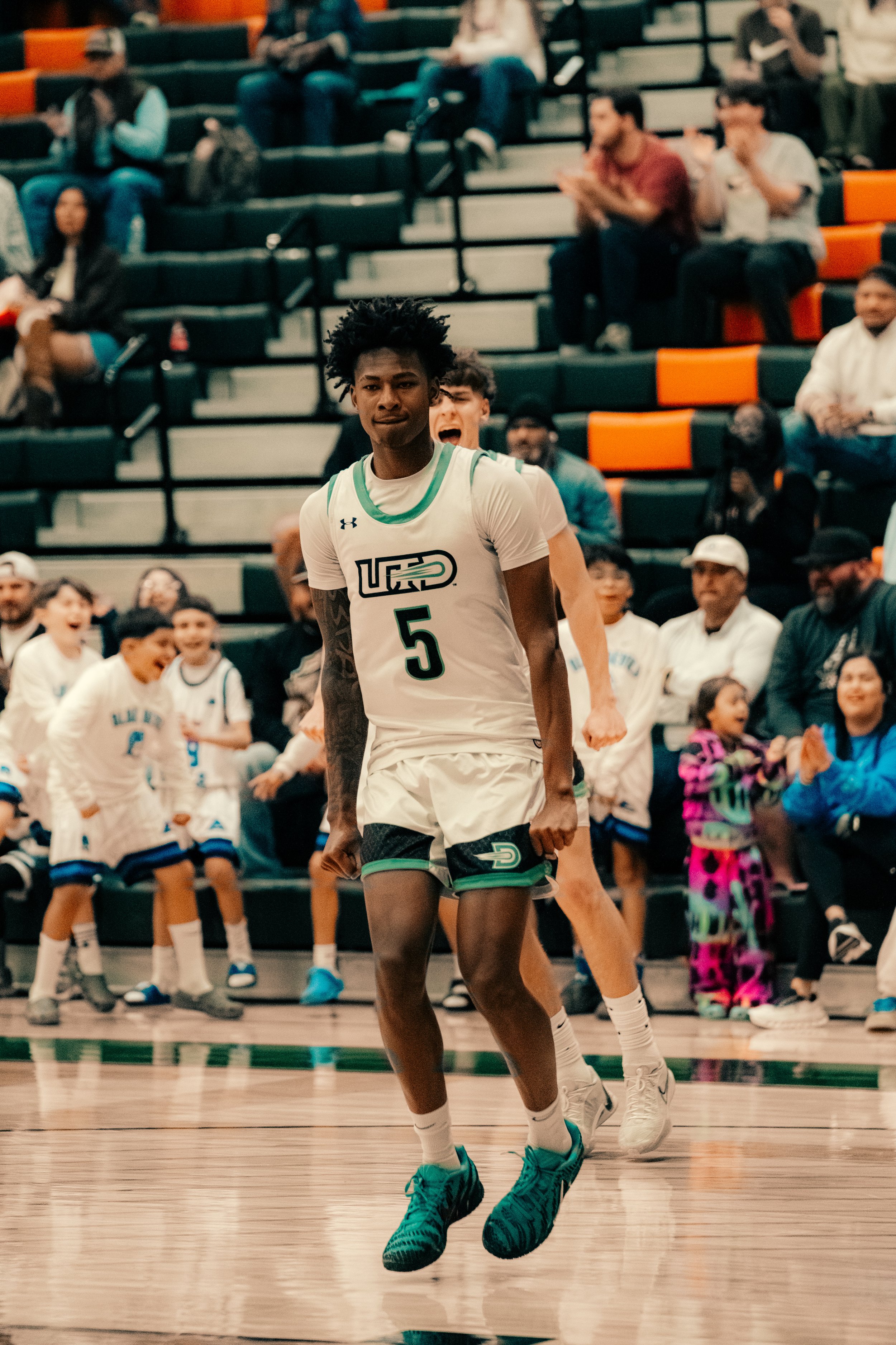 A basketball player in a white jersey with the number 5 is running on a court during a game. The background shows spectators and other players, some of whom are children and others are adults, watching and cheering.