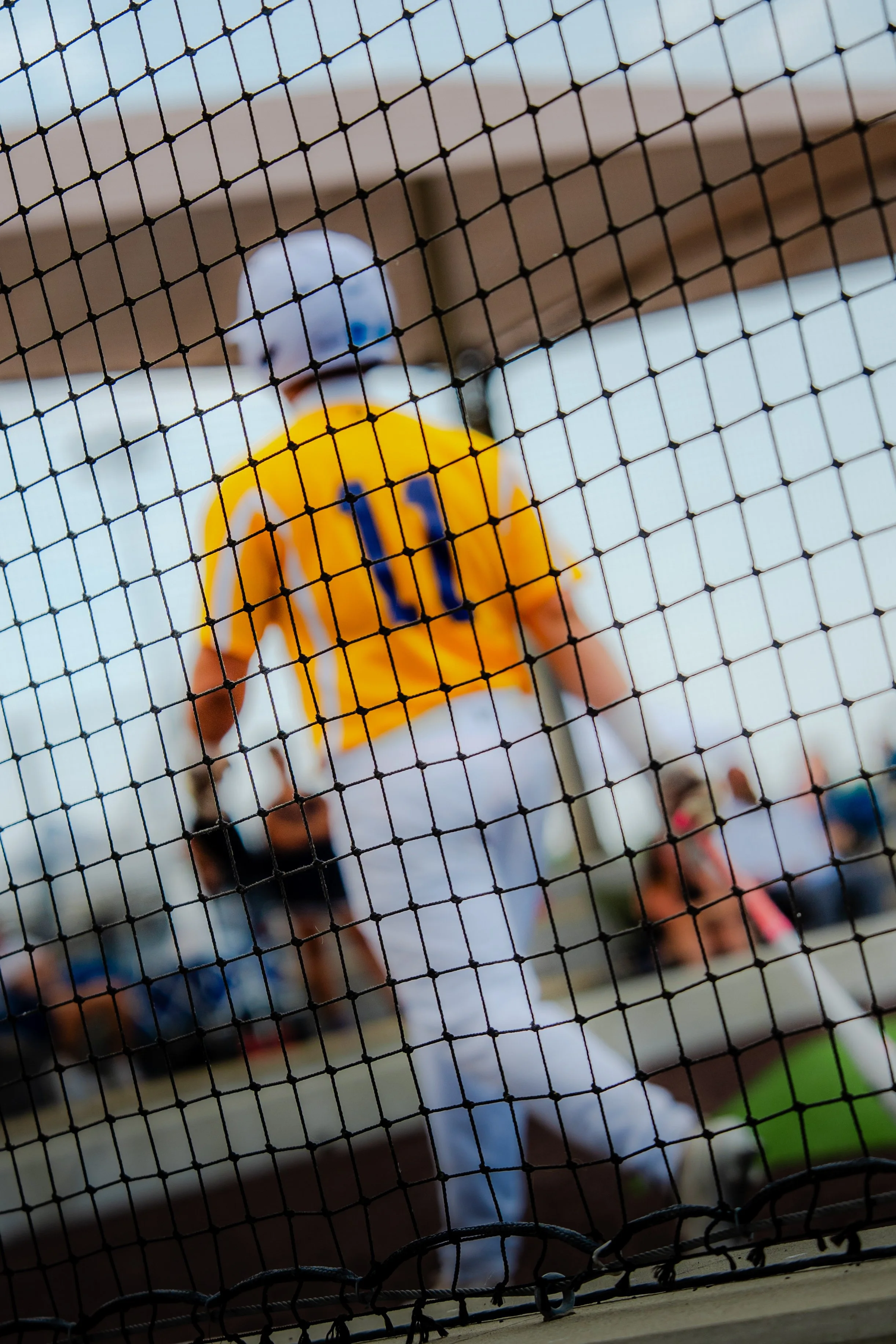 A person in a yellow sports jersey with the number 10 on it is behind a black wire fence. They are wearing a white baseball cap and are outdoors on a sunny day.