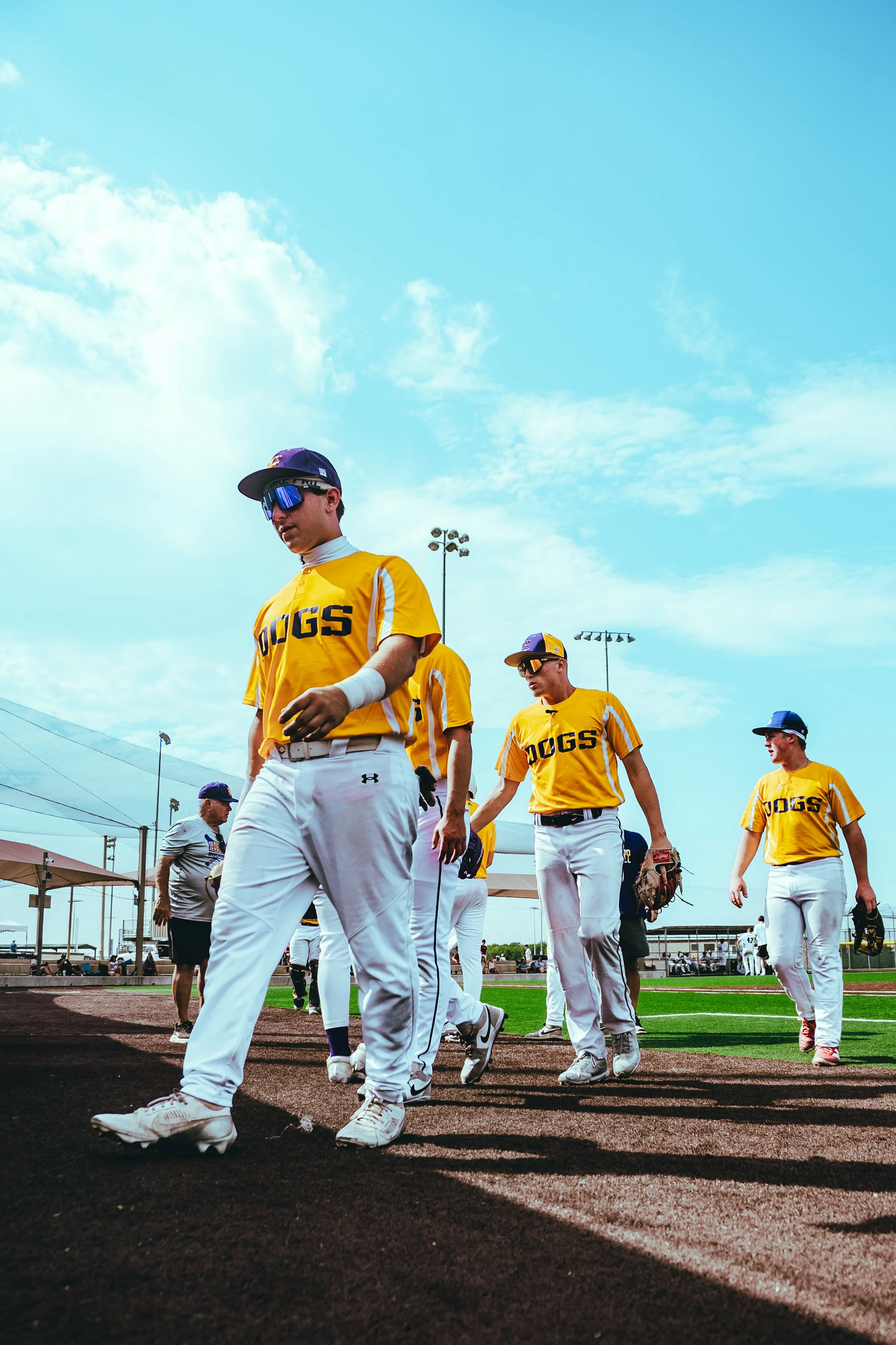 Baseball players in yellow jerseys walking on the field during a game, with a clear blue sky and stadium lights in the background.