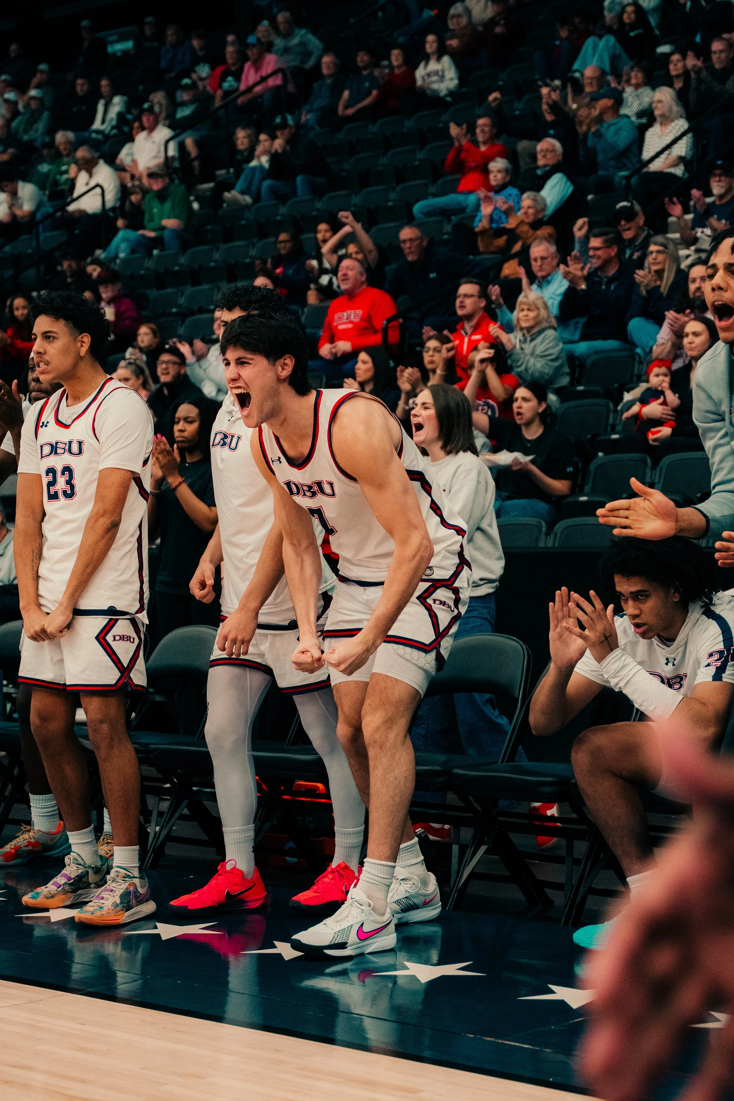 A basketball game scene with players in white jerseys standing on the sideline, fans in the background cheering and clapping in a stadium.