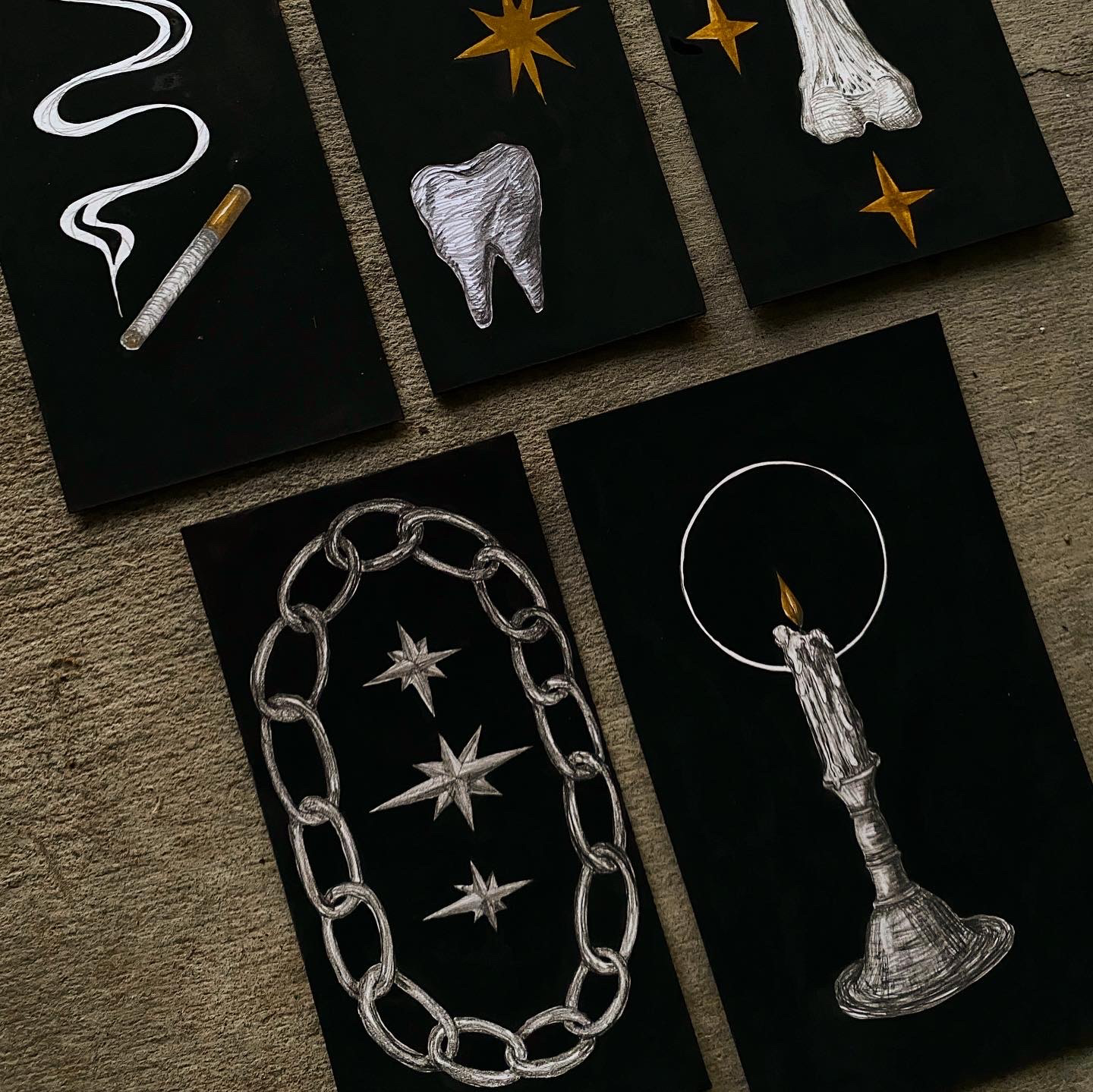 Collection of five black cards, each with silver or gold artistic illustrations of objects such as a tooth, a mouth, a chain, a candle, and stars, with some featuring gold accents.