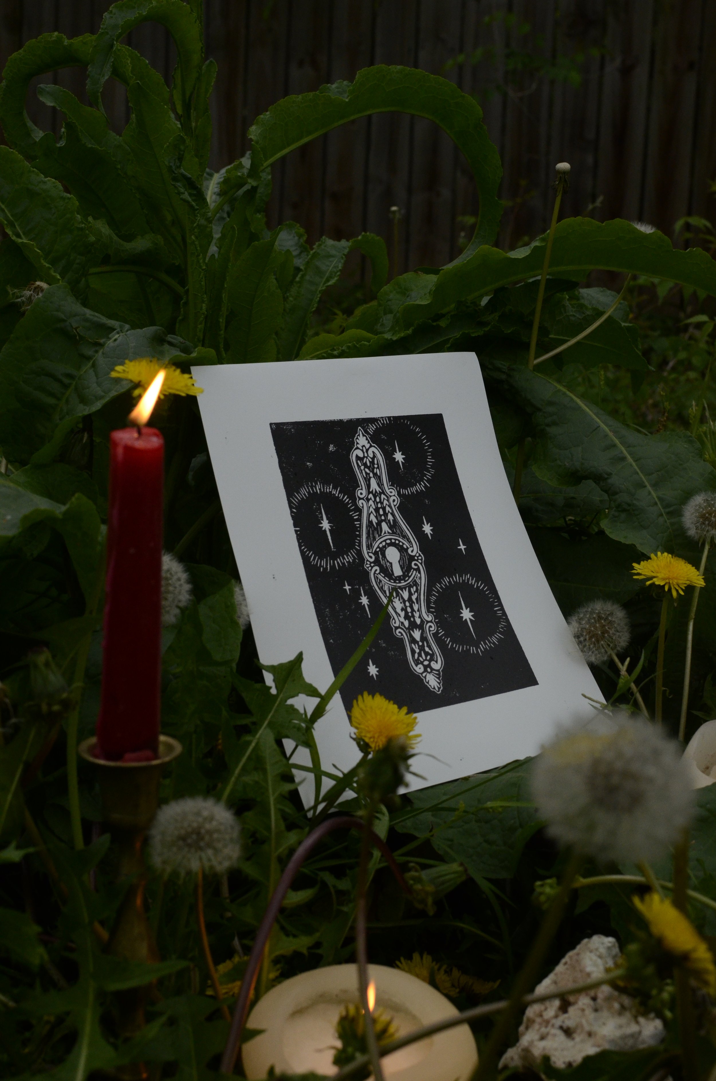 A small candle flame surrounded by flowers, a card featuring a mystical illustration, and a dark background with a wooden fence.