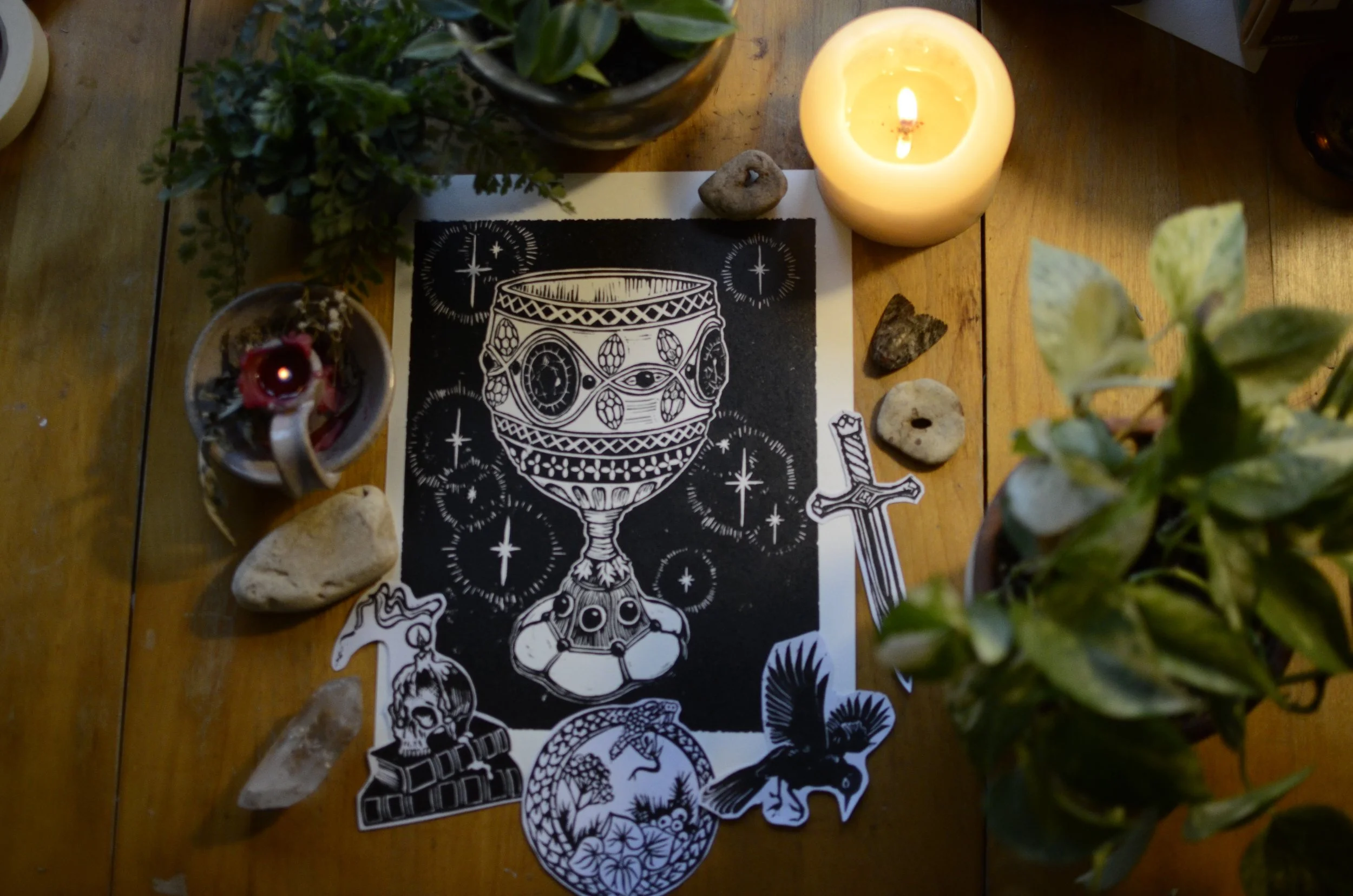 A black and white sticker sheet featuring various illustrations of a goblet, skeletal skulls, hand, snake, owl, and plants, placed on a wooden table among candles, stones, and plants.