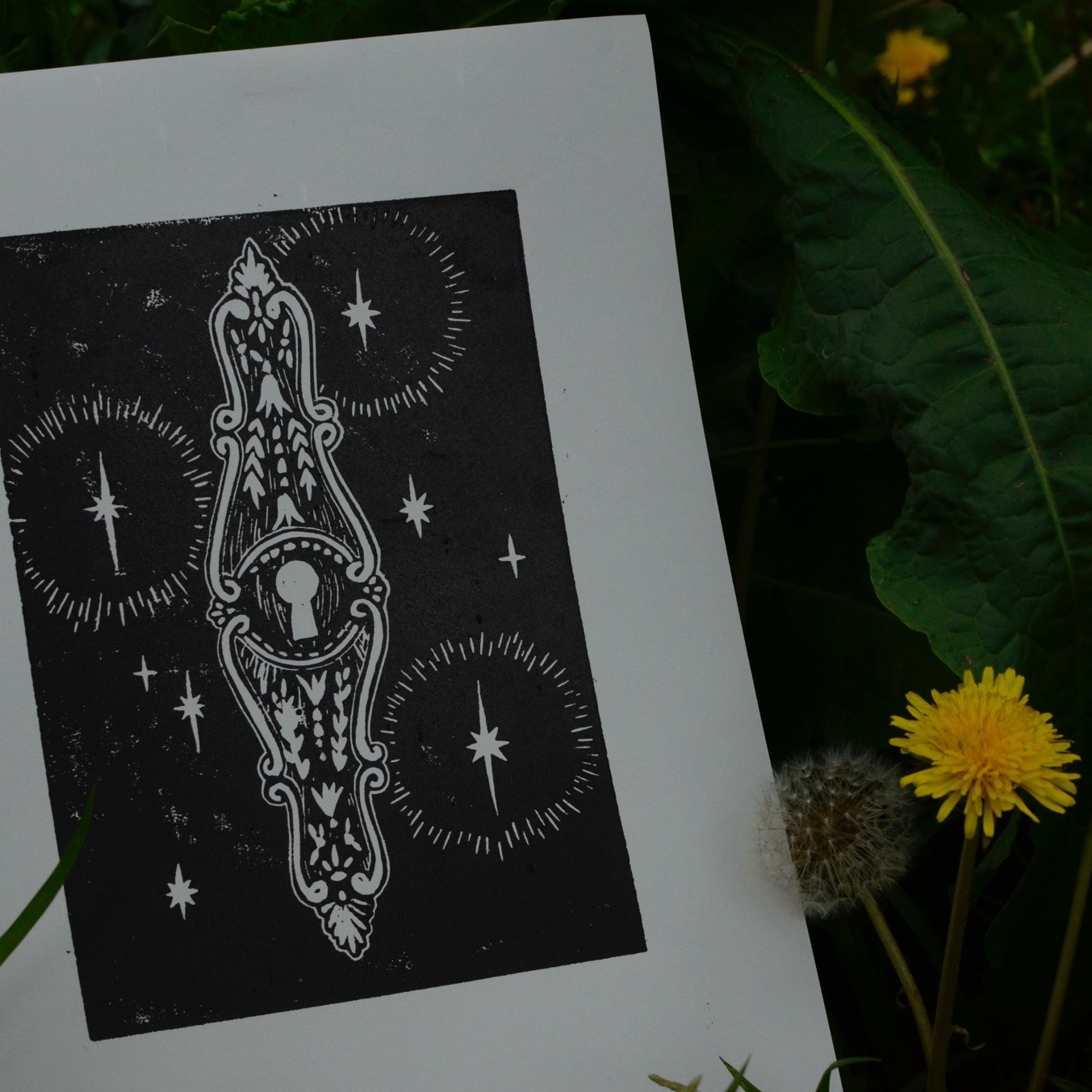 A black and white tarot card with intricate designs and star motifs, placed on green leaves with a yellow dandelion flower and a small dandelion puff nearby.