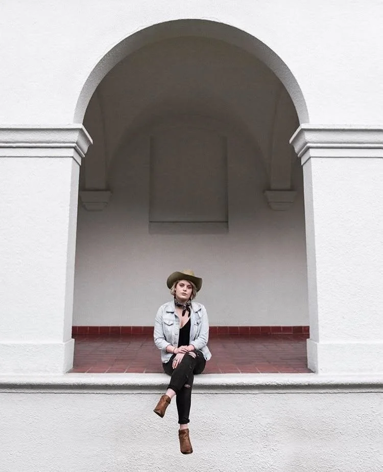 A woman sitting on a ledge in front of a white building with an arched opening, wearing a hat, denim jacket, black pants, and boots.