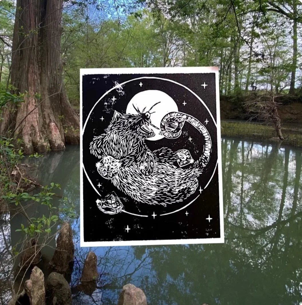 A black and white digital illustration of a monstrous unicorn with a twisted horn, surrounded by stars, moons, and other cosmic elements. The illustration appears to be a sticker or printed artwork placed in front of a natural scene with a tree, wate