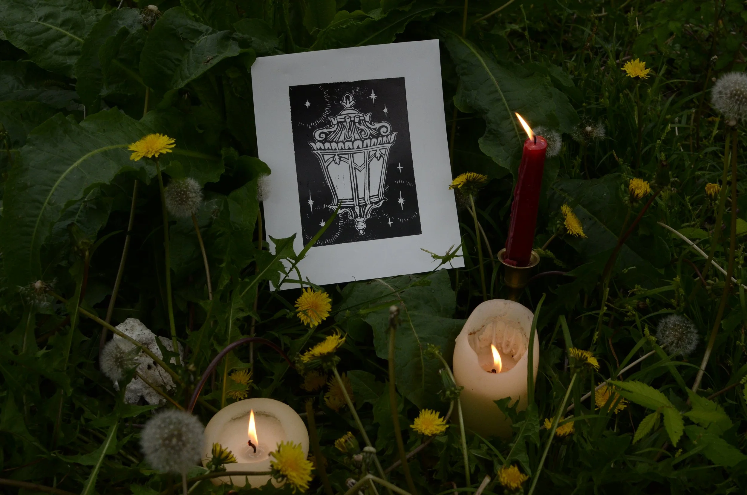 A memorial display with a black and white illustration of a lantern, surrounded by yellow flowers, dandelions, and lit candles, set among green foliage.