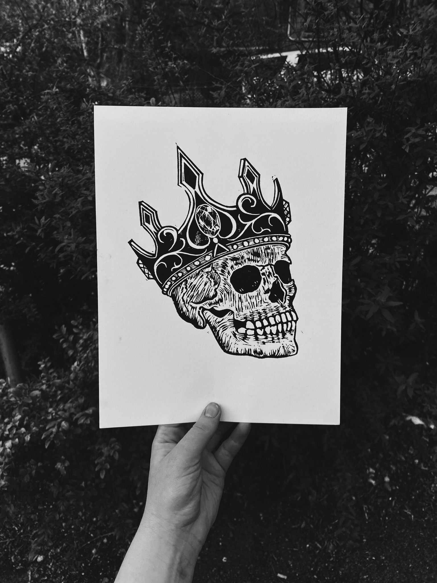 A black and white drawing of a human skull wearing a crown with a black gemstone on the forehead, held by a person's hand in front of bushes.