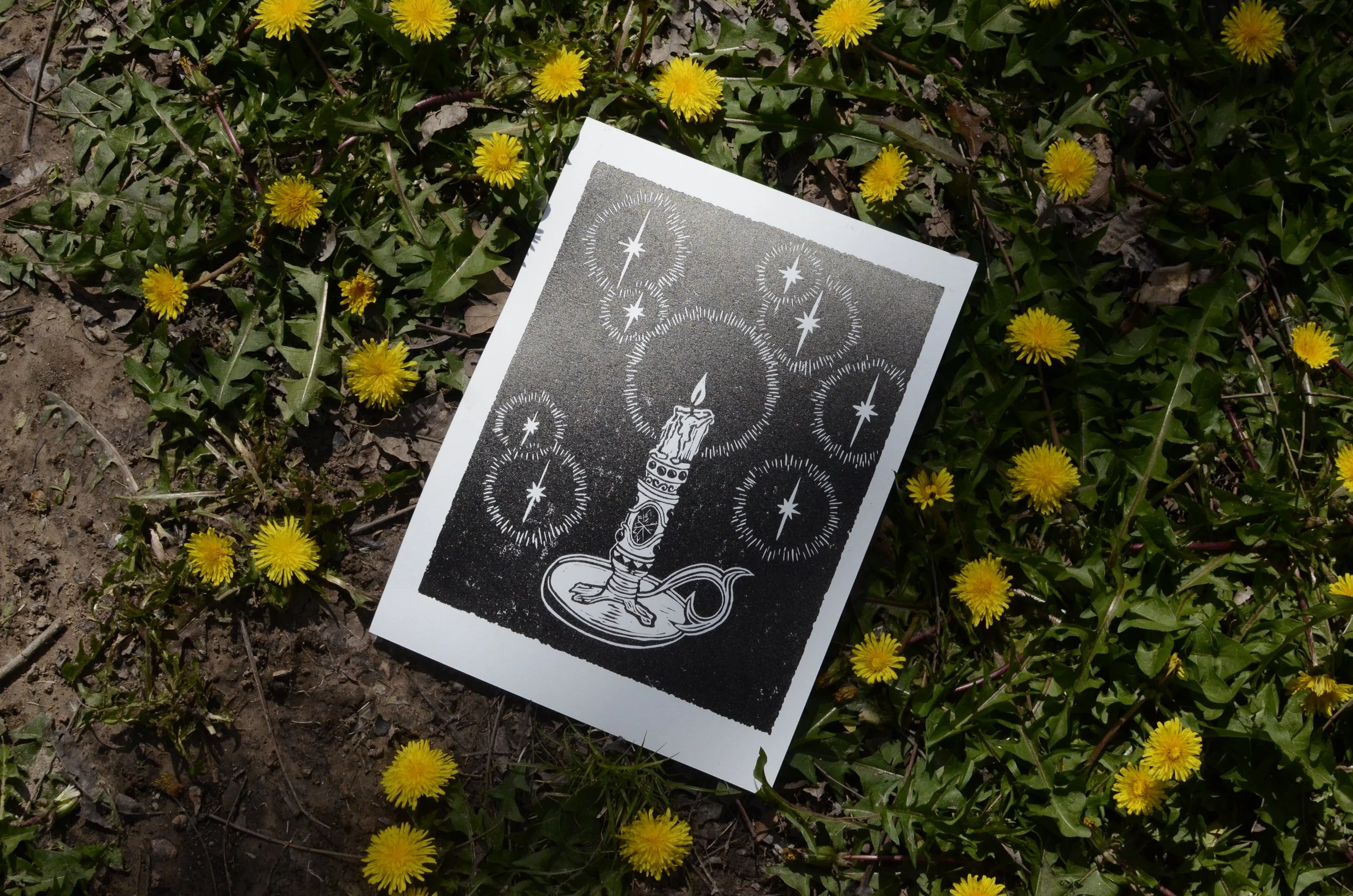 A tarot card with a candle illustration surrounded by star patterns lying on ground among yellow dandelion flowers and green leaves.