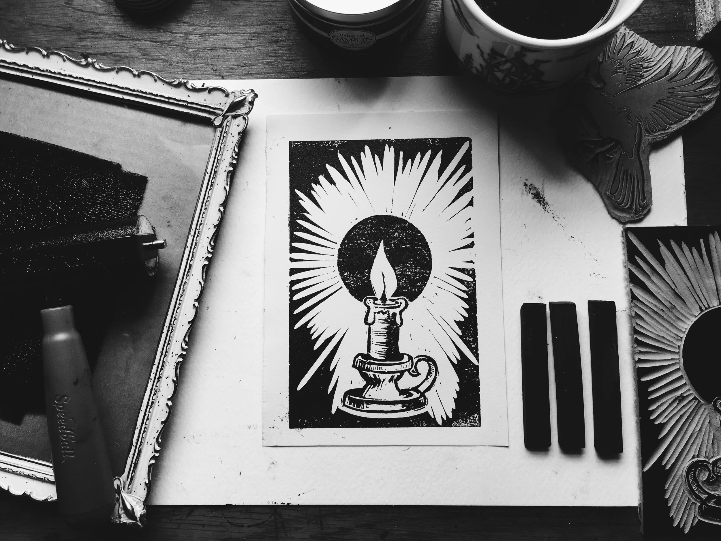 A black and white photo of a desk with art supplies and artwork. There is a framed piece, a candle labeled 'Relax Lap Camellia,' a cup of coffee, and a printed image of a candle with a radiant background. Also present are a butterfly-shaped decoratio