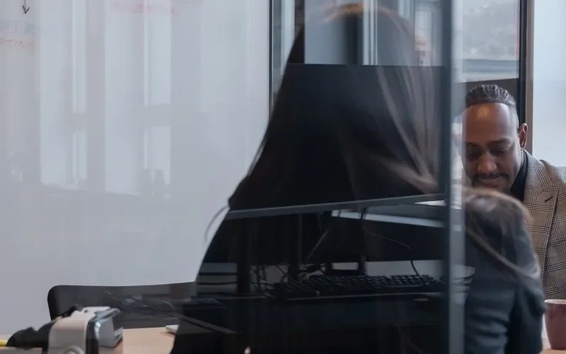 Two people in a modern office, engaged in a conversation behind a glass partition, with a computer monitor on the desk.