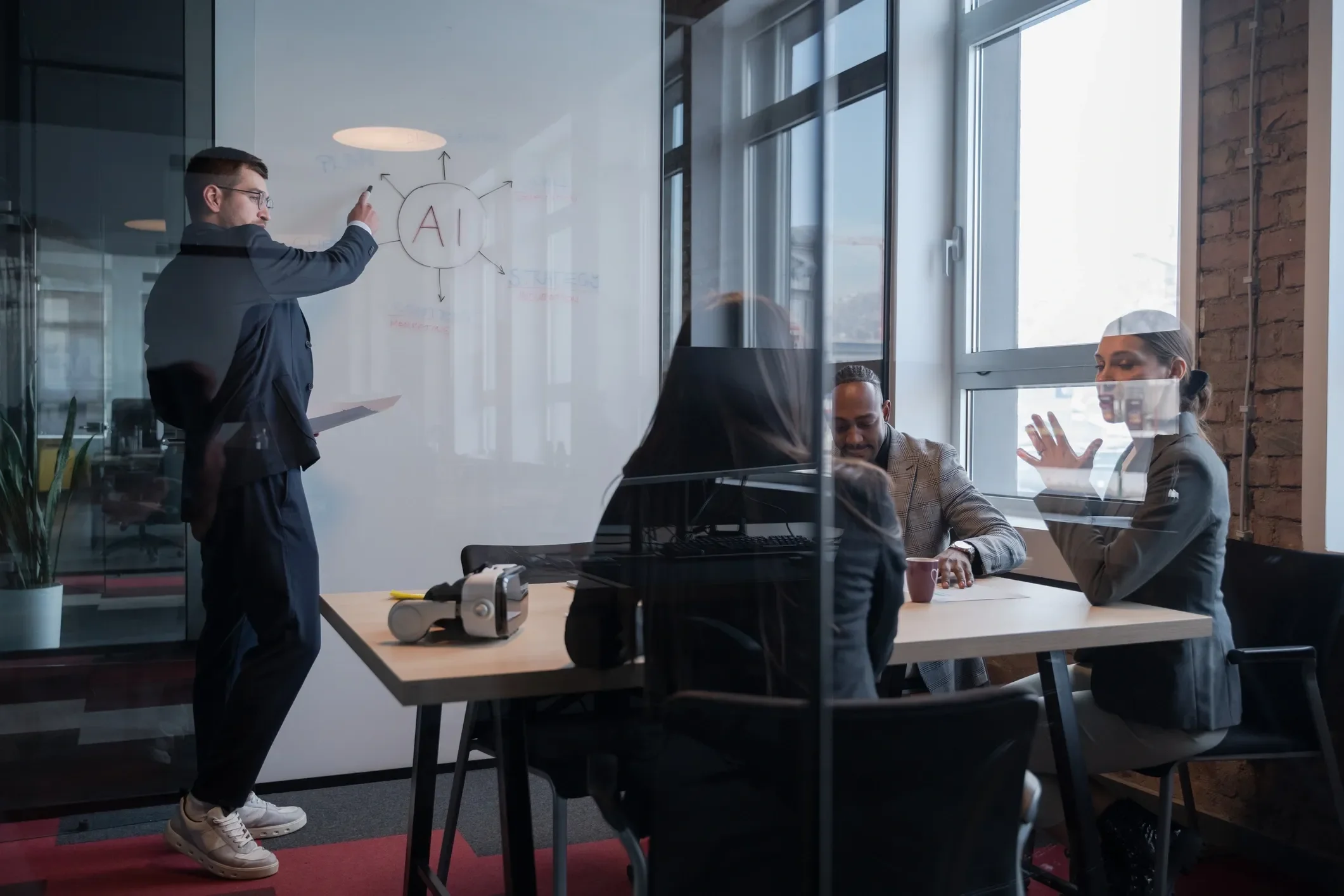 A man in a suit and glasses is writing 'AI' on a whiteboard in a modern office. A group of three people, including a woman with brown hair, are sitting at a table behind a glass wall, engaged in a discussion. The office has large windows and exposed brick walls.